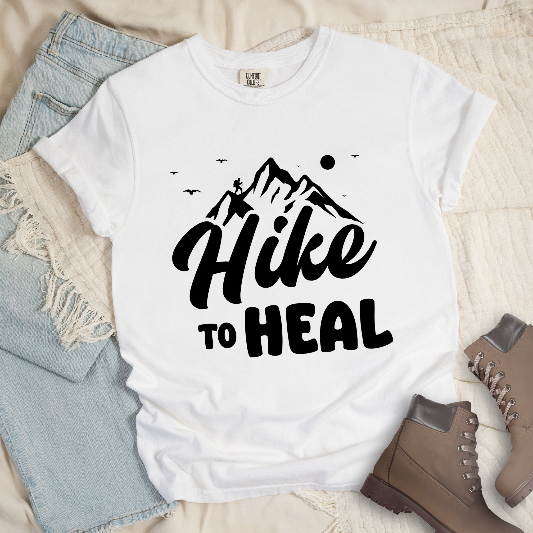 White graphic t-shirt with 'Hike to Heal' text in black with mountain and bird design, styled with light wash jeans and brown hiking boots