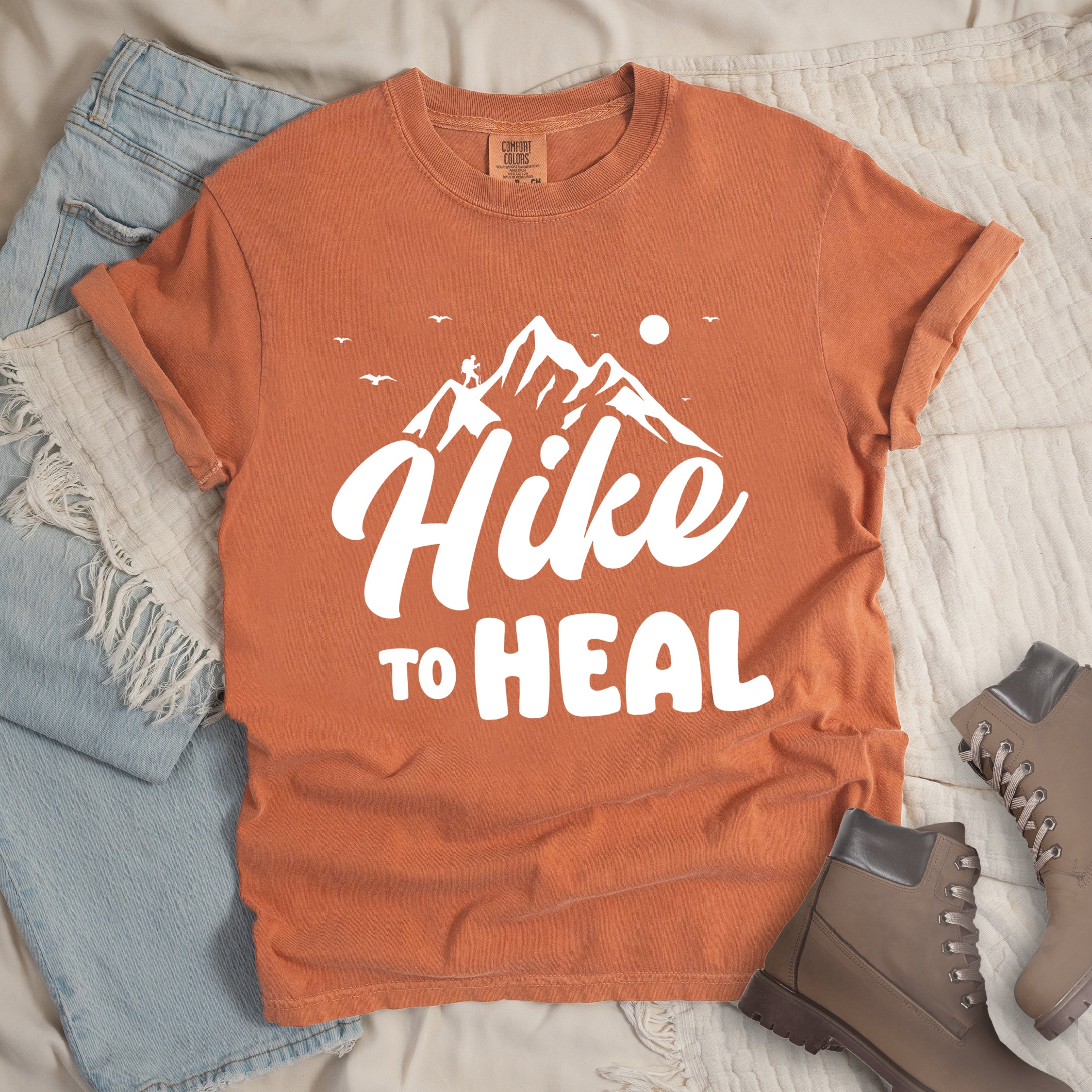 Yam (Brown) graphic t-shirt with 'Hike to Heal' text in white with mountain and bird design, styled with light wash jeans and brown hiking boots