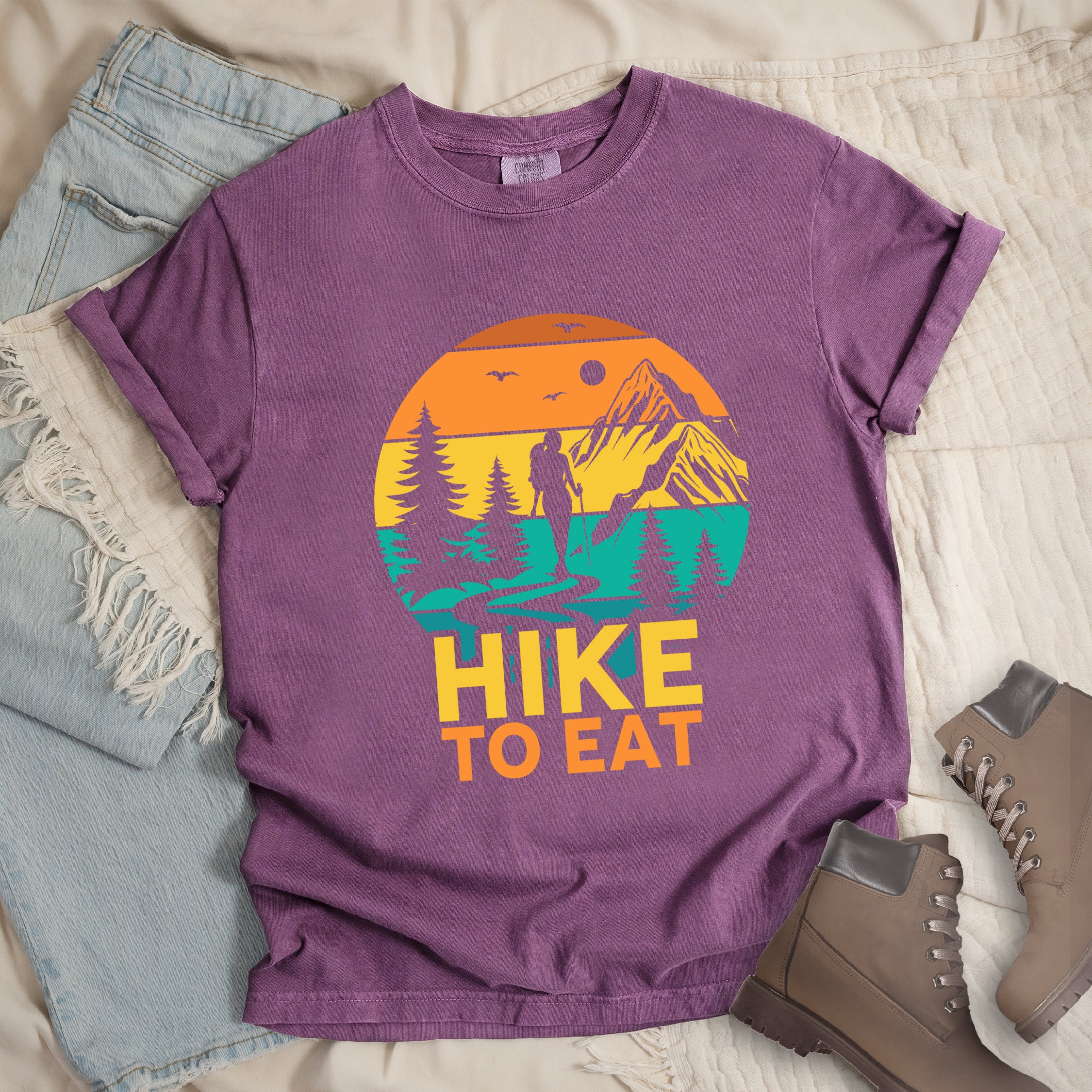 Berry (Dark Violet) graphic t-shirt with 'Hike to Eat' text and colorful sunset mountain scene featuring hiker silhouette among pine trees, styled with light wash jeans and brown hiking boots