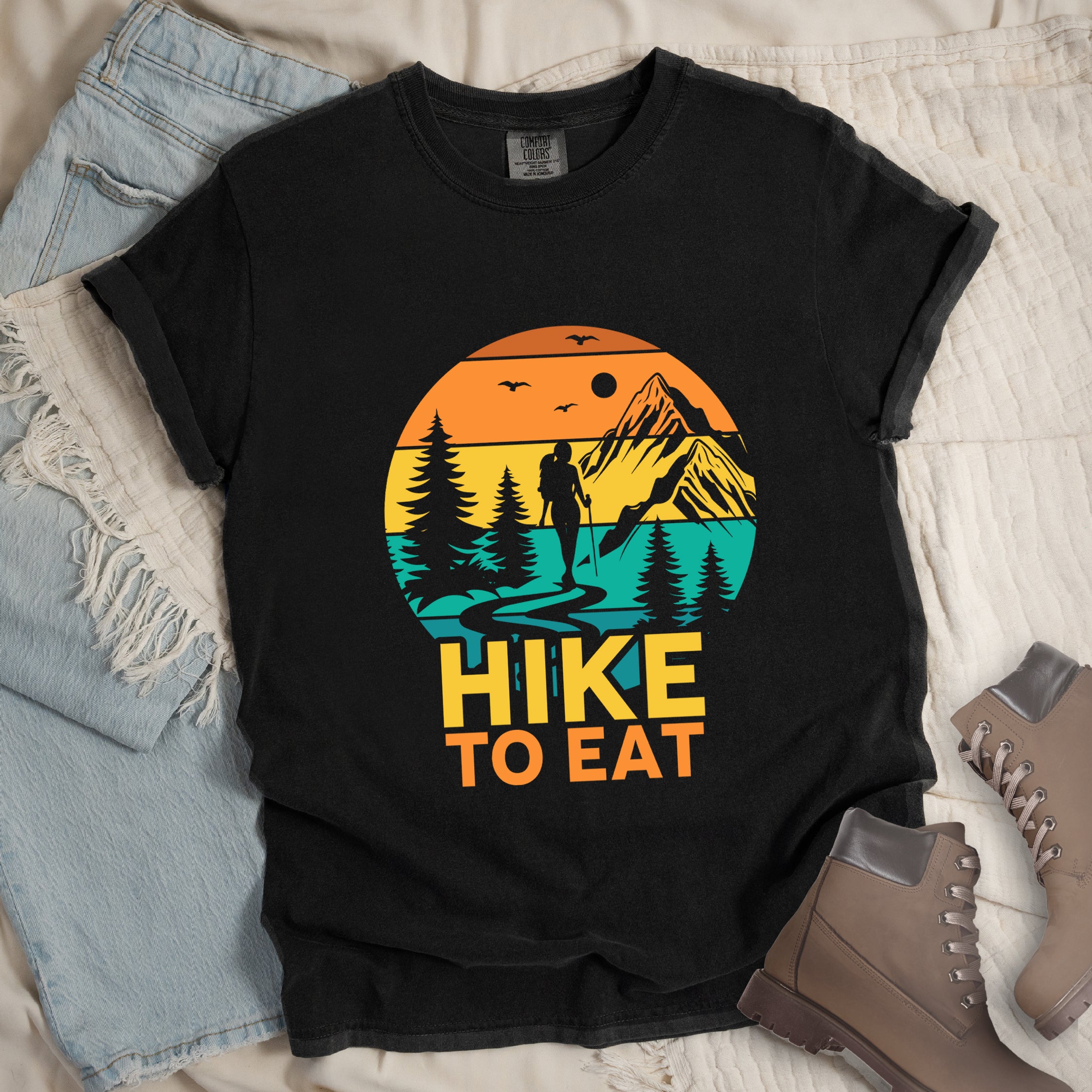 Black graphic t-shirt with 'Hike to Eat' text and colorful sunset mountain scene featuring hiker silhouette among pine trees, styled with light wash jeans and brown hiking boots
