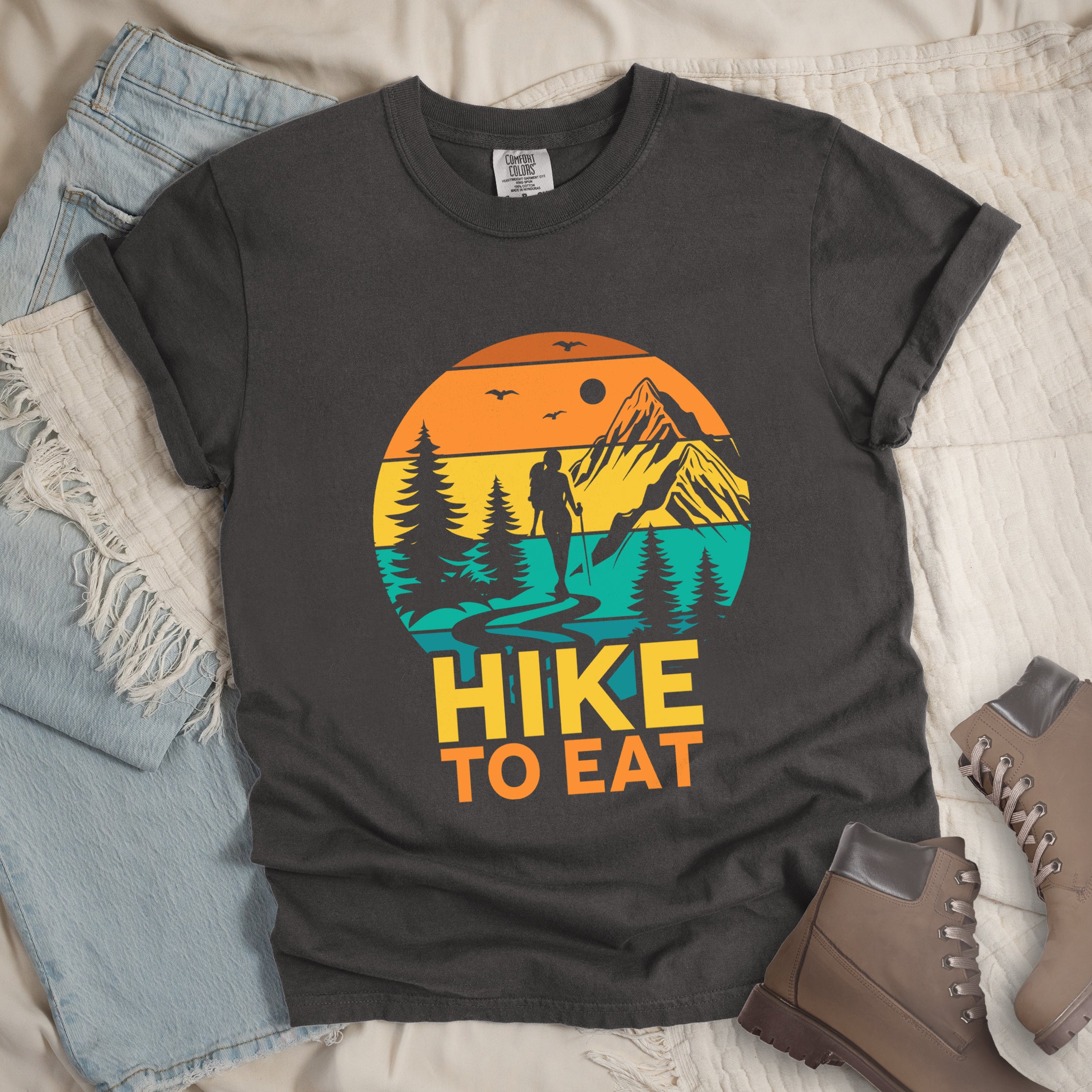 Pepper (Dark Gray) graphic t-shirt with 'Hike to Eat' text and colorful sunset mountain scene featuring hiker silhouette among pine trees, styled with light wash jeans and brown hiking boots