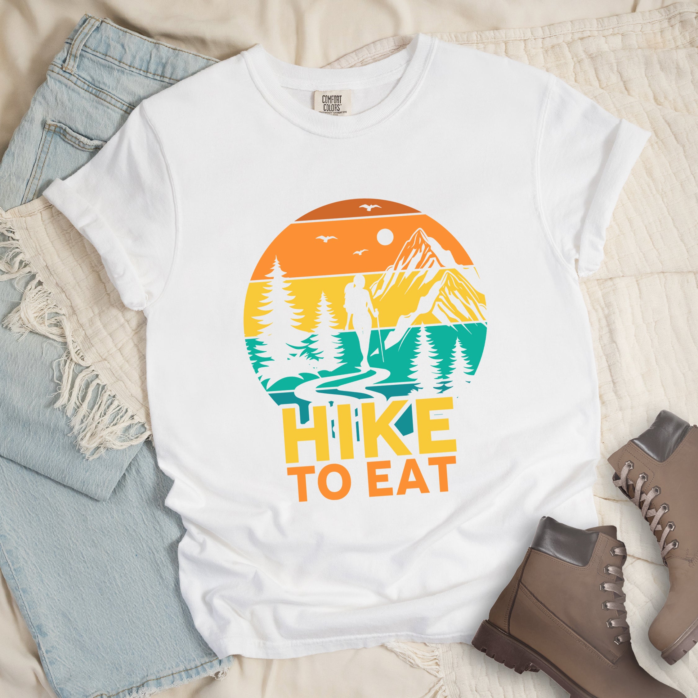 White graphic t-shirt with 'Hike to Eat' text and colorful sunset mountain scene featuring hiker silhouette among pine trees, styled with light wash jeans and brown hiking boots