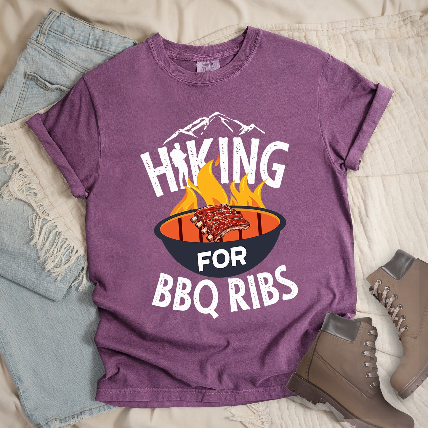 Berry (Dark Violet) t-shirt with white distressed text reading "HIKING FOR BBQ RIBS". Mountain silhouettes appear above the word "HIKING". The letter "I" in "HIKING" features a hiker silhouette. Between "HIKING" and "BBQ RIBS" is an illustration of a grill with flames and a rack of ribs cooking. The word "FOR" appears in white on the grill. Features a classic crew neck and casual rolled sleeves, styled alongside light wash denim and brown hiking boots.
