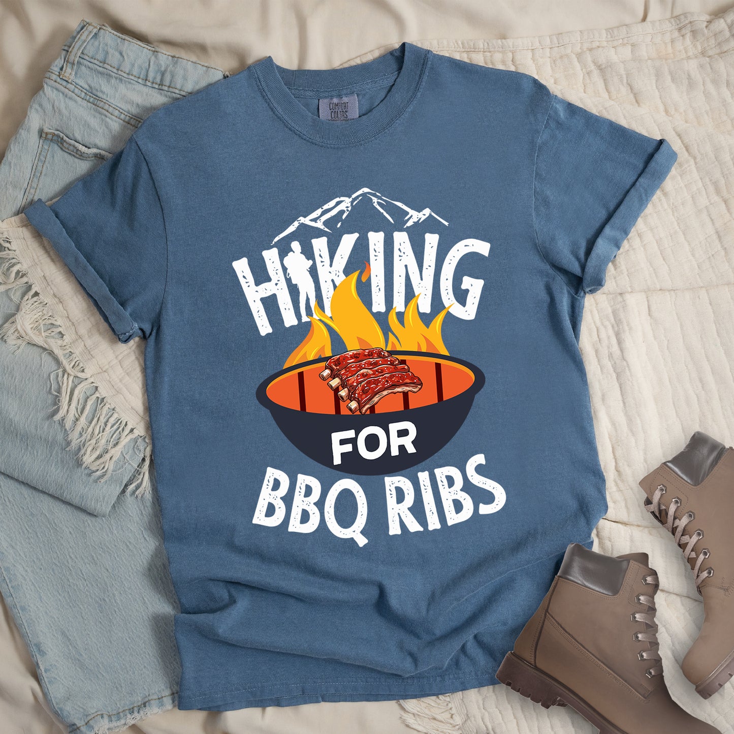 Blue Jean (Denim Blue) t-shirt with white distressed text reading "HIKING FOR BBQ RIBS". Mountain silhouettes appear above the word "HIKING". The letter "I" in "HIKING" features a hiker silhouette. Between "HIKING" and "BBQ RIBS" is an illustration of a grill with flames and a rack of ribs cooking. The word "FOR" appears in white on the grill. Features a classic crew neck and casual rolled sleeves, styled alongside light wash denim and brown hiking boots.