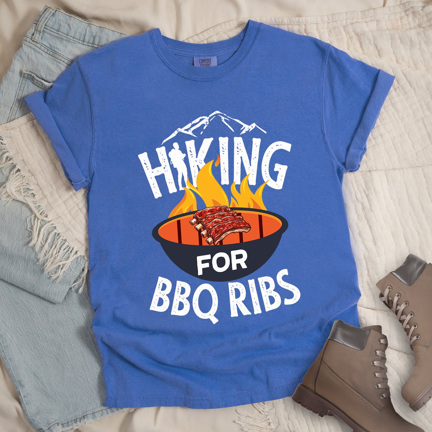 Flo Blue (Bright Blue) t-shirt with white distressed text reading "HIKING FOR BBQ RIBS". Mountain silhouettes appear above the word "HIKING". The letter "I" in "HIKING" features a hiker silhouette. Between "HIKING" and "BBQ RIBS" is an illustration of a grill with flames and a rack of ribs cooking. The word "FOR" appears in white on the grill. Features a classic crew neck and casual rolled sleeves, styled alongside light wash denim and brown hiking boots.