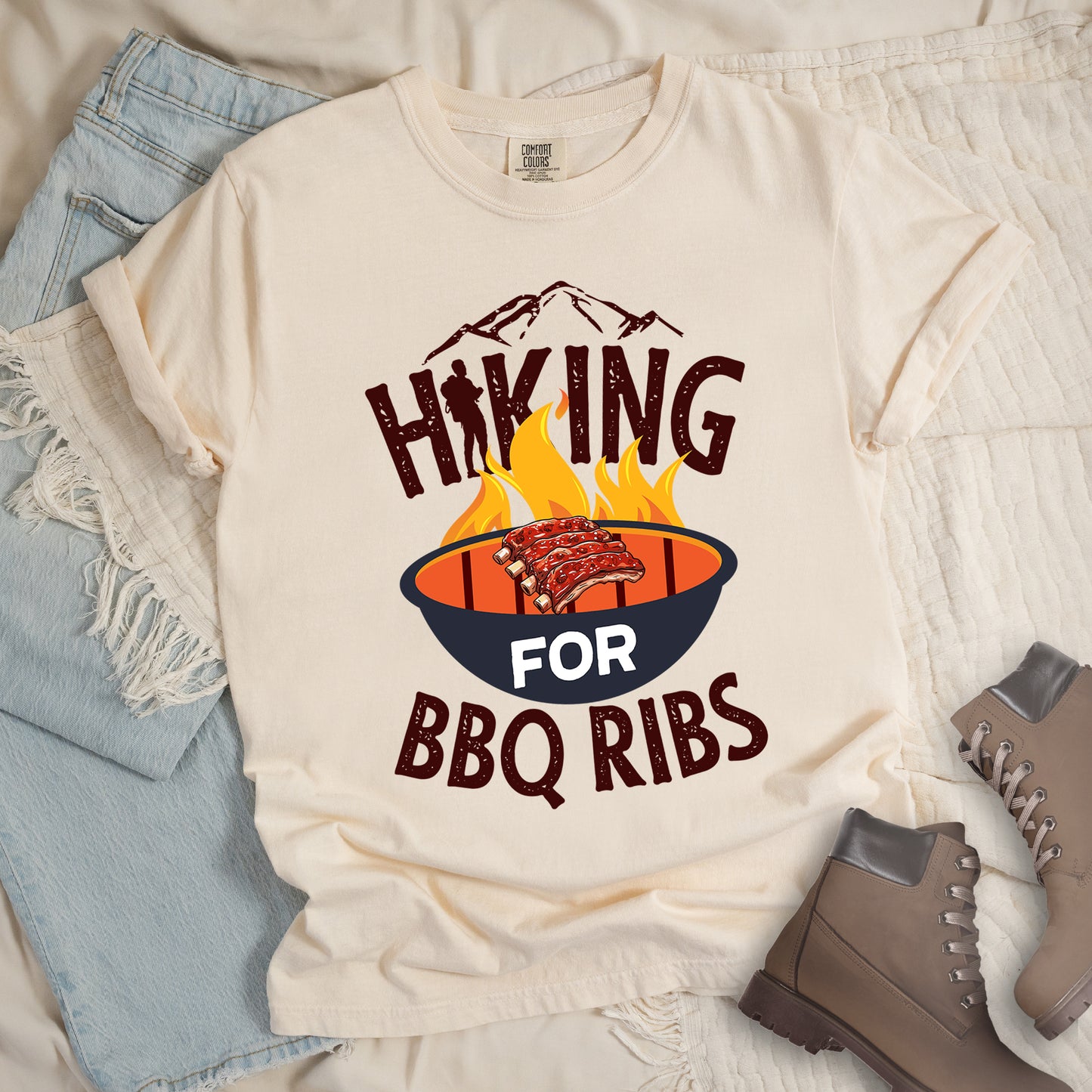 Ivory (Cream White) t-shirt with dark brown distressed text reading "HIKING FOR BBQ RIBS". Mountain silhouettes appear above the word "HIKING". The letter "I" in "HIKING" features a hiker silhouette. Between "HIKING" and "BBQ RIBS" is an illustration of a grill with flames and a rack of ribs cooking. The word "FOR" appears in white on the grill. Features a classic crew neck and casual rolled sleeves, styled alongside light wash denim and brown hiking boots.