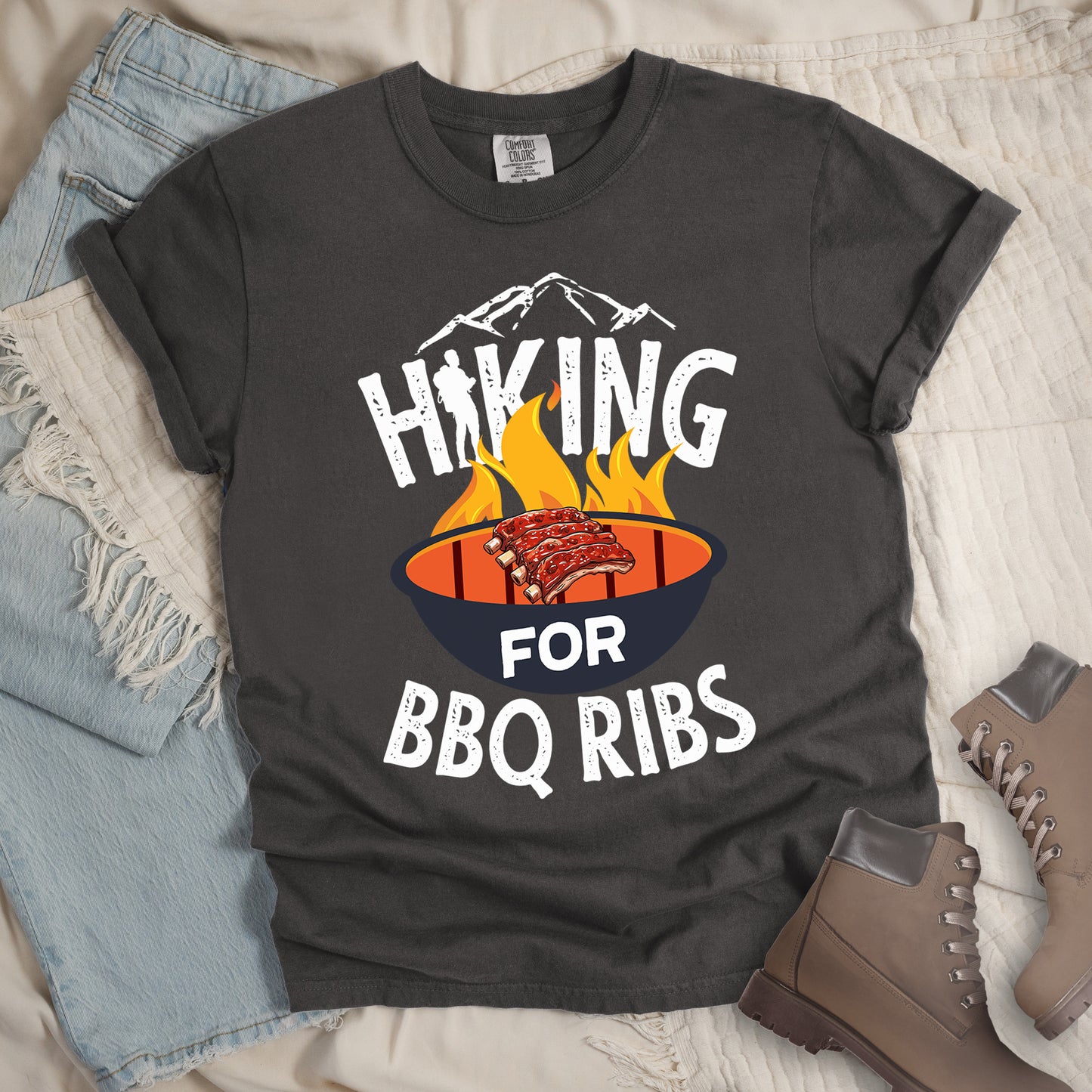 Pepper (Dark Gray) t-shirt with white distressed text reading "HIKING FOR BBQ RIBS". Mountain silhouettes appear above the word "HIKING". The letter "I" in "HIKING" features a hiker silhouette. Between "HIKING" and "BBQ RIBS" is an illustration of a grill with flames and a rack of ribs cooking. The word "FOR" appears in white on the grill. Features a classic crew neck and casual rolled sleeves, styled alongside light wash denim and brown hiking boots.
