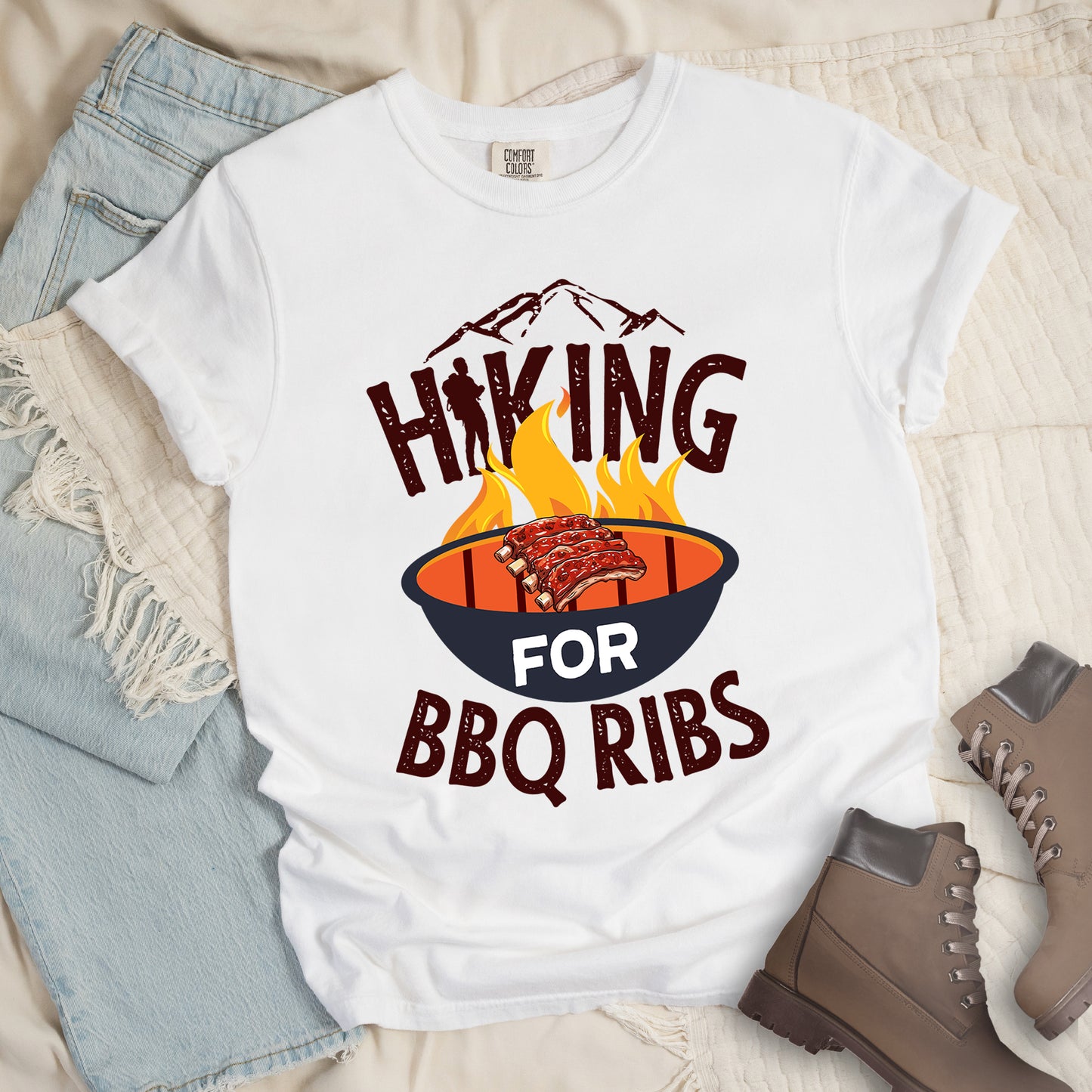 White t-shirt with dark brown distressed text reading "HIKING FOR BBQ RIBS". Mountain silhouettes appear above the word "HIKING". The letter "I" in "HIKING" features a hiker silhouette. Between "HIKING" and "BBQ RIBS" is an illustration of a grill with flames and a rack of ribs cooking. The word "FOR" appears in white on the grill. Features a classic crew neck and casual rolled sleeves, styled alongside light wash denim and brown hiking boots.