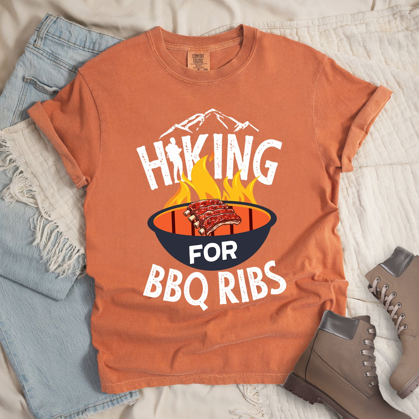 Yam (Brown) t-shirt with white distressed text reading "HIKING FOR BBQ RIBS". Mountain silhouettes appear above the word "HIKING". The letter "I" in "HIKING" features a hiker silhouette. Between "HIKING" and "BBQ RIBS" is an illustration of a grill with flames and a rack of ribs cooking. The word "FOR" appears in white on the grill. Features a classic crew neck and casual rolled sleeves, styled alongside light wash denim and brown hiking boots.