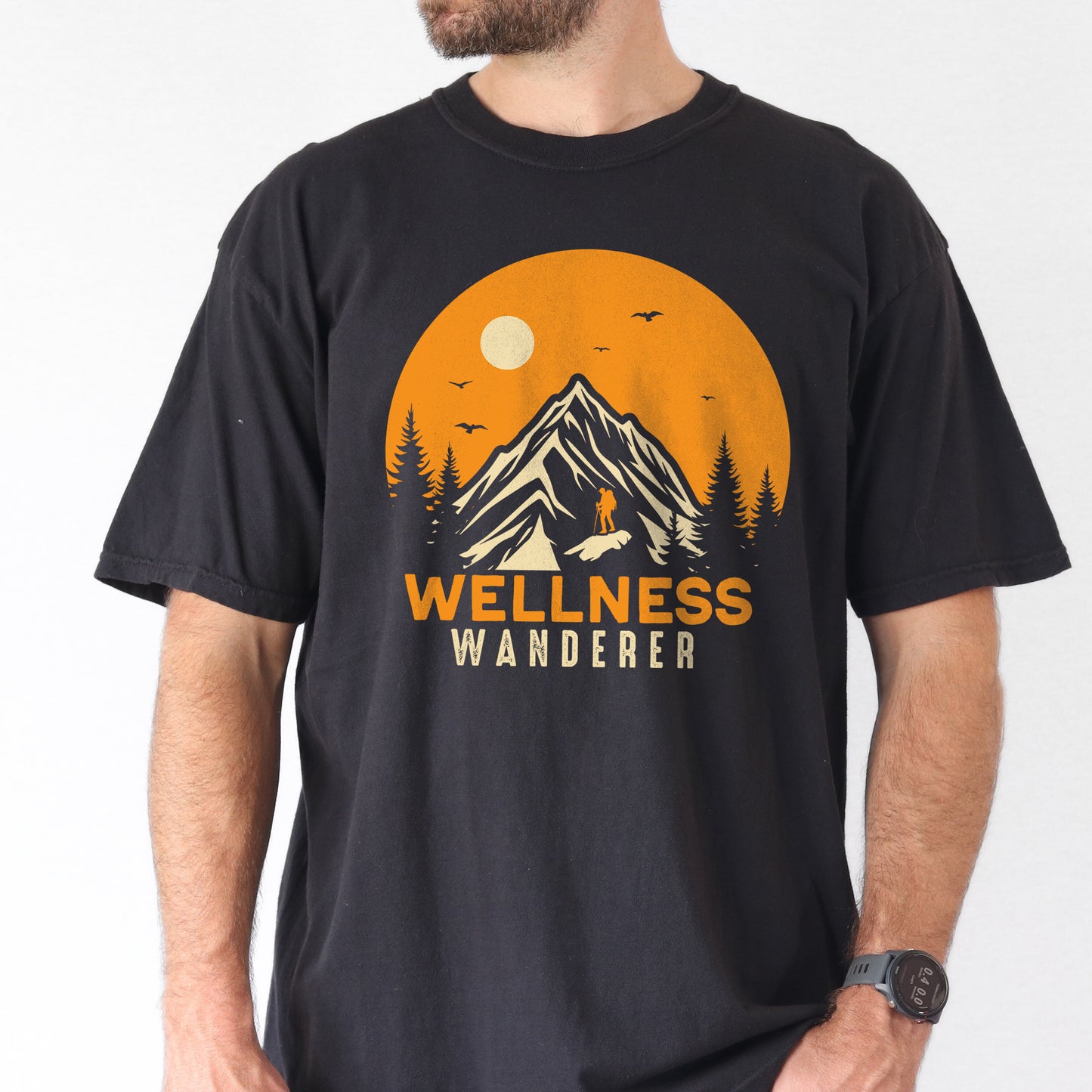 Black graphic t-shirt with 'Wellness Wanderer' text in orange lettering featuring circular sunset design with mountains, pine trees, hiker silhouette, flying birds, and sun.