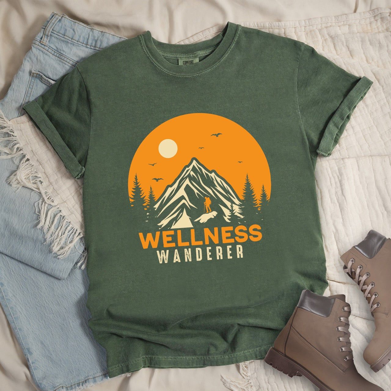 Moss Wellness lifestyle vintage tee featuring mountain peak landscape with character silhouette at summit orange sunset circle flying birds pine trees and bold Wellness Wanderer typography on Comfort Colors shirt for nature lovers seeking balance