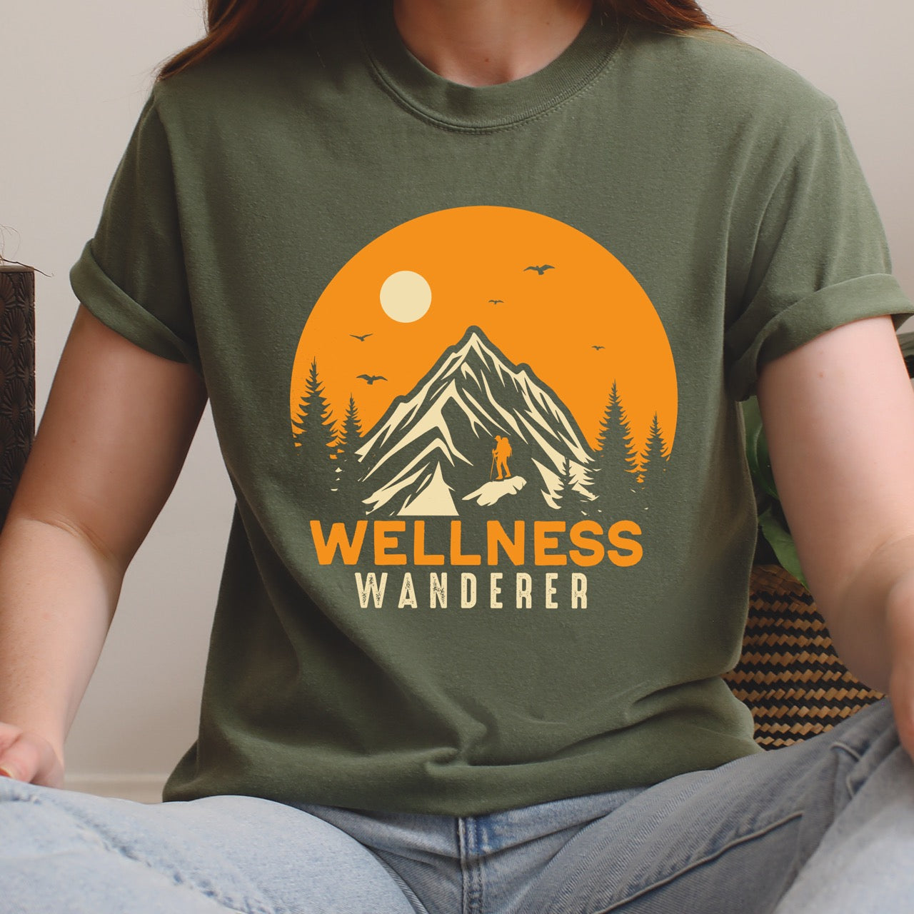 Woman wearing moss Wellness lifestyle vintage tee featuring mountain peak landscape with character silhouette at summit orange sunset circle flying birds pine trees and bold Wellness Wanderer typography on Comfort Colors shirt for nature lovers seeking balance