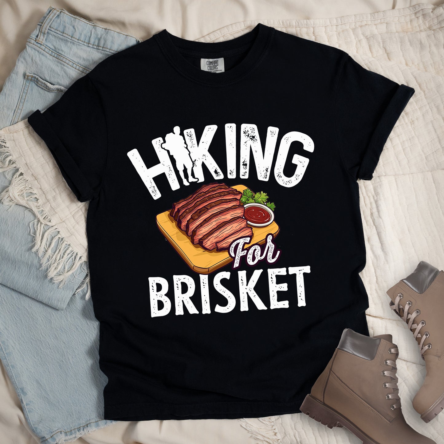 Black t-shirt with white distressed text reading "HIKING For BRISKET". The letter "I" in "HIKING" features a hiker silhouette with backpack. Between the words is an illustration of sliced brisket on a wooden cutting board with a small side of sauce and garnish. The word "For" appears in script lettering. Features a classic crew neck and casual rolled sleeves, styled alongside light wash denim and brown hiking boots.