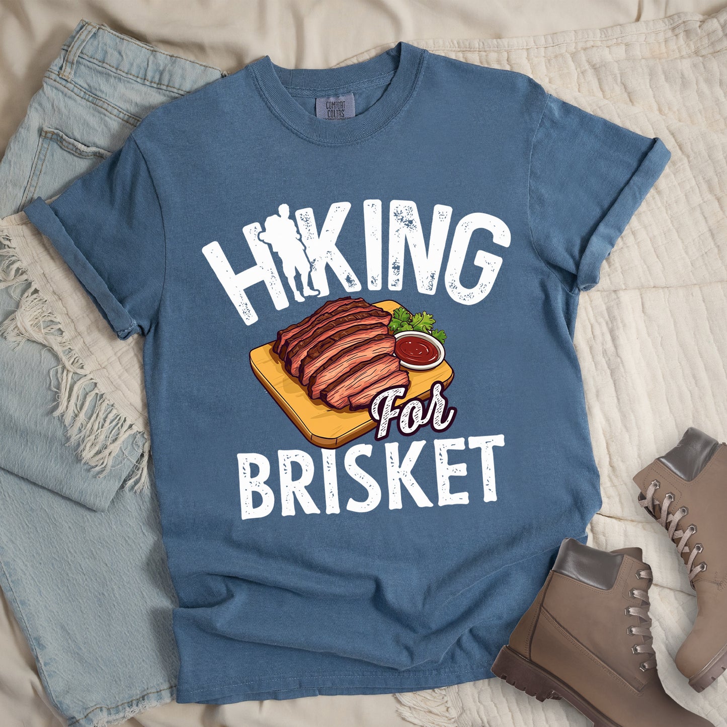 Blue Jean (Denim Blue) t-shirt with white distressed text reading "HIKING For BRISKET". The letter "I" in "HIKING" features a hiker silhouette with backpack. Between the words is an illustration of sliced brisket on a wooden cutting board with a small side of sauce and garnish. The word "For" appears in script lettering. Features a classic crew neck and casual rolled sleeves, styled alongside light wash denim and brown hiking boots.