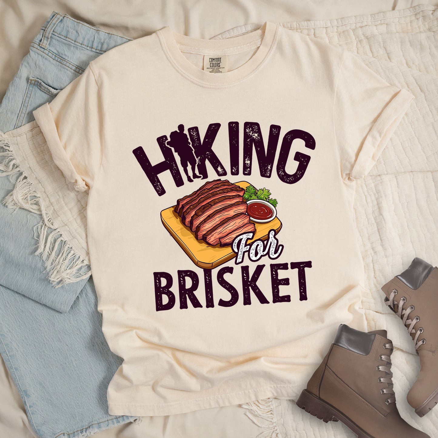 Ivory (Cream White) t-shirt with dark brown distressed text reading "HIKING For BRISKET". The letter "I" in "HIKING" features a hiker silhouette with backpack. Between the words is an illustration of sliced brisket on a wooden cutting board with a small side of sauce and garnish. The word "For" appears in script lettering. Features a classic crew neck and casual rolled sleeves, styled alongside light wash denim and brown hiking boots.