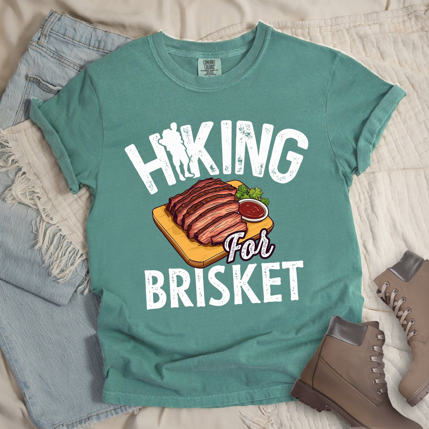 Light Green t-shirt with white distressed text reading "HIKING For BRISKET". The letter "I" in "HIKING" features a hiker silhouette with backpack. Between the words is an illustration of sliced brisket on a wooden cutting board with a small side of sauce and garnish. The word "For" appears in script lettering. Features a classic crew neck and casual rolled sleeves, styled alongside light wash denim and brown hiking boots.