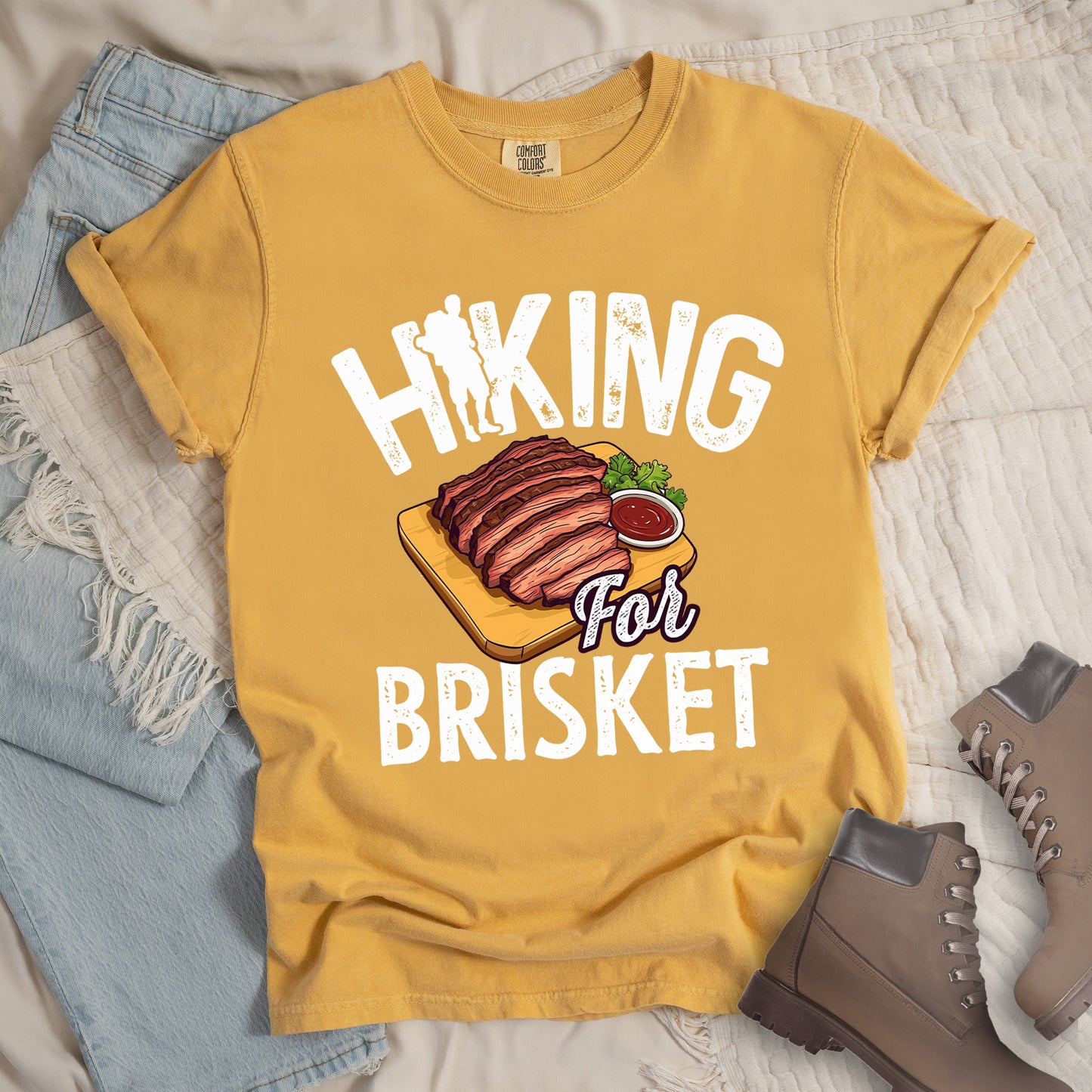 Mustard (Yellow) t-shirt with white distressed text reading "HIKING For BRISKET". The letter "I" in "HIKING" features a hiker silhouette with backpack. Between the words is an illustration of sliced brisket on a wooden cutting board with a small side of sauce and garnish. The word "For" appears in script lettering. Features a classic crew neck and casual rolled sleeves, styled alongside light wash denim and brown hiking boots.
