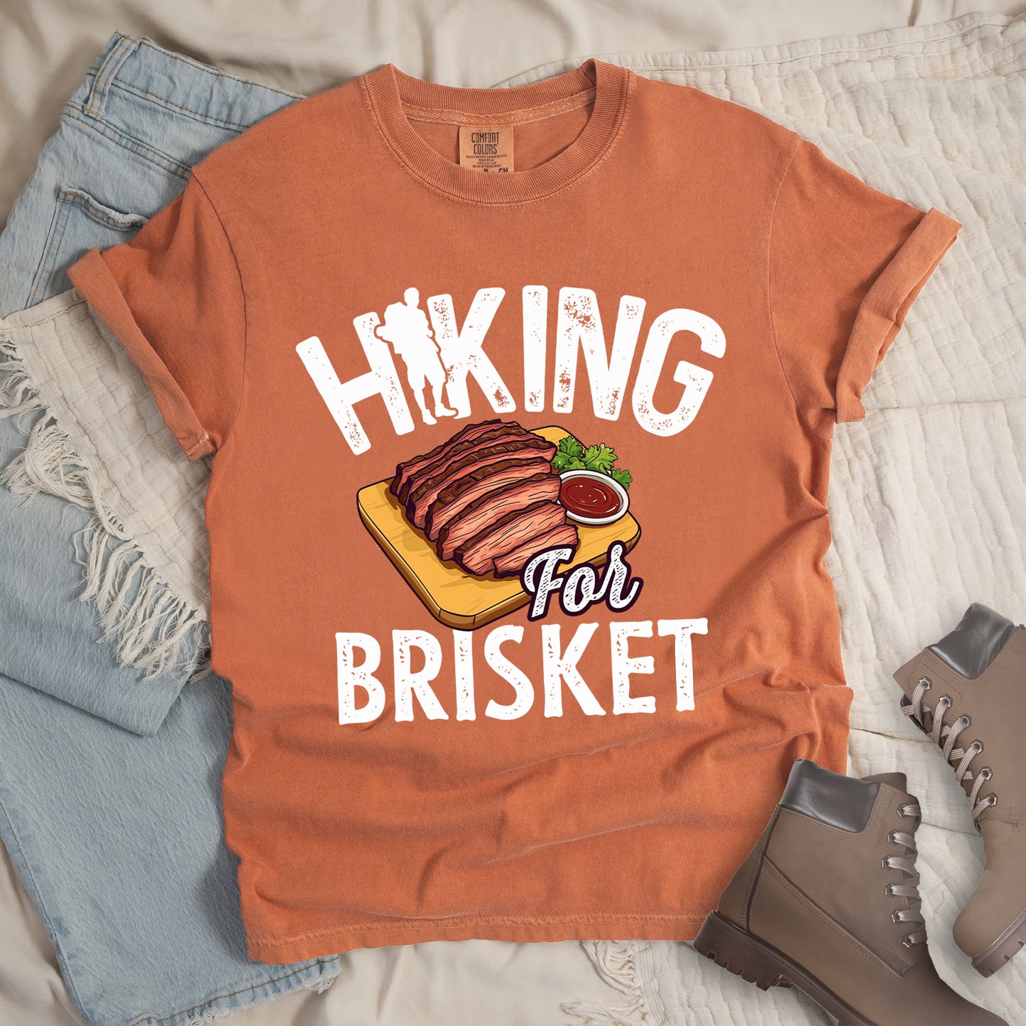 Yam (Brown) t-shirt with white distressed text reading "HIKING For BRISKET". The letter "I" in "HIKING" features a hiker silhouette with backpack. Between the words is an illustration of sliced brisket on a wooden cutting board with a small side of sauce and garnish. The word "For" appears in script lettering. Features a classic crew neck and casual rolled sleeves, styled alongside light wash denim and brown hiking boots.