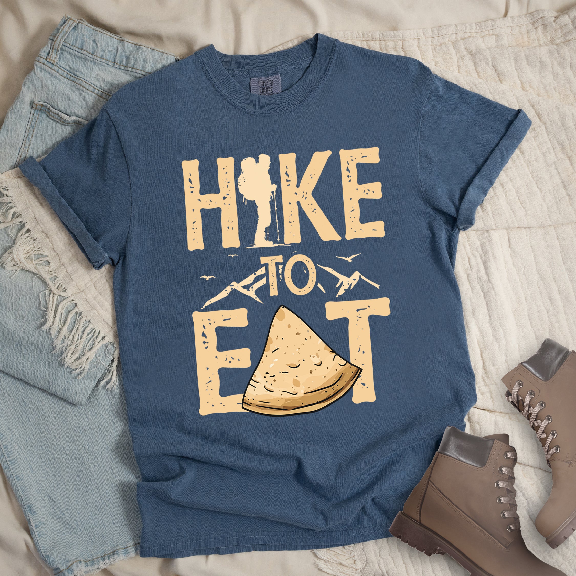 Blue Jean (Denim Blue) t-shirt with text "HIKE TO EAT". "HIKE" appears in large beige letters at the top with a hiker silhouette as letter "I", "TO" in beige text with small mountain outlines on either side in the middle, and "EAT" in large beige letters at the bottom with a triangular slice of flatbread or pastry (tortilla) replacing the letter "A". Small flying birds appear near "TO". Features a classic crew neck and casual rolled sleeves, styled alongside light wash denim and brown hiking boots.