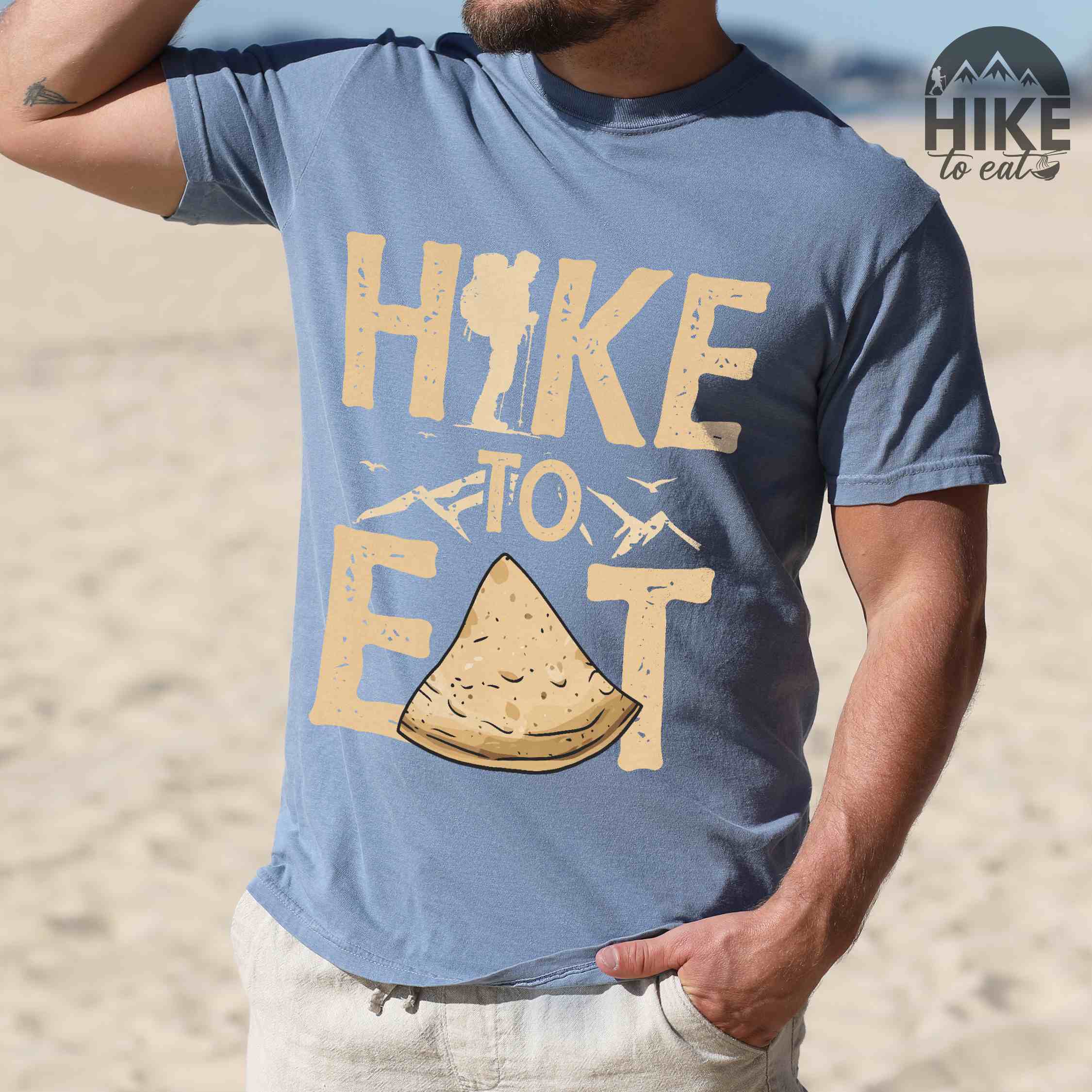 Man wearing blue jean "Hike to Eat" colored statement tee with bold typography, mountain, illustrated tortilla, perfect for tortilla lovers.