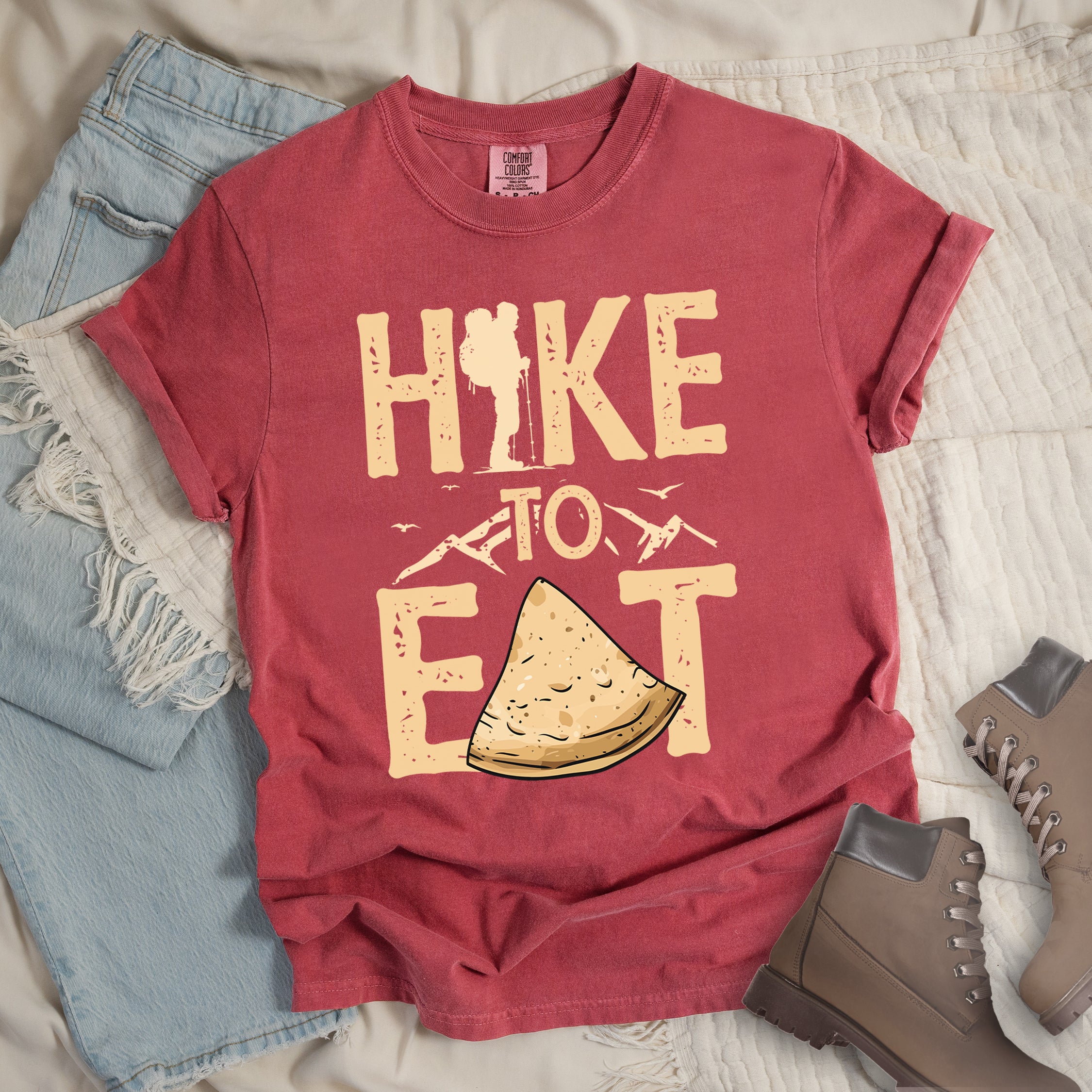 Crimson (Dark Pink)t-shirt with text "HIKE TO EAT". "HIKE" appears in large beige letters at the top with a hiker silhouette as letter "I", "TO" in beige text with small mountain outlines on either side in the middle, and "EAT" in large beige letters at the bottom with a triangular slice of flatbread or pastry (tortilla) replacing the letter "A". Small flying birds appear near "TO". Features a classic crew neck and casual rolled sleeves, styled alongside light wash denim and brown hiking boots.