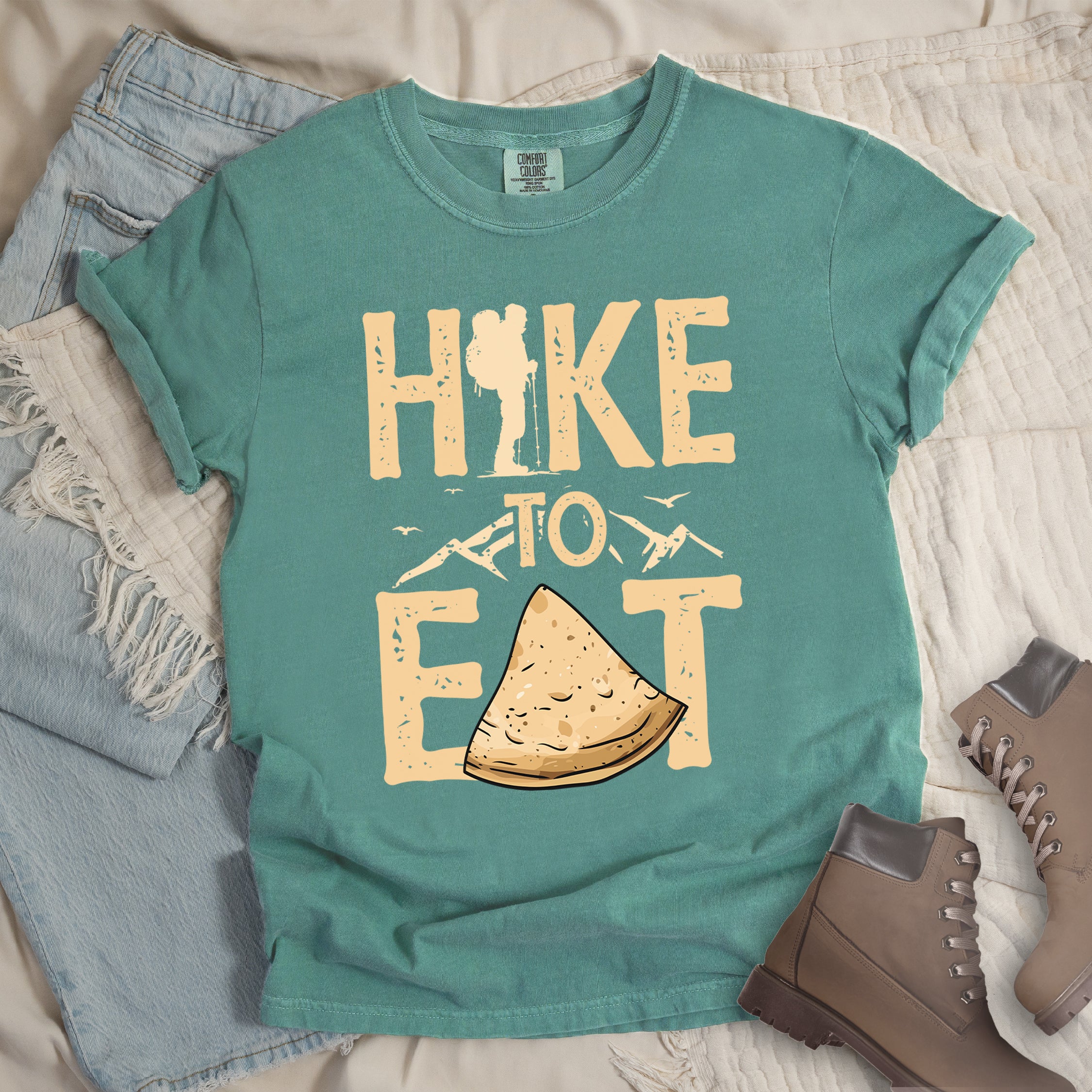 Light Green t-shirt with text "HIKE TO EAT". "HIKE" appears in large beige letters at the top with a hiker silhouette as letter "I", "TO" in beige text with small mountain outlines on either side in the middle, and "EAT" in large beige letters at the bottom with a triangular slice of flatbread or pastry (tortilla) replacing the letter "A". Small flying birds appear near "TO". Features a classic crew neck and casual rolled sleeves, styled alongside light wash denim and brown hiking boots.