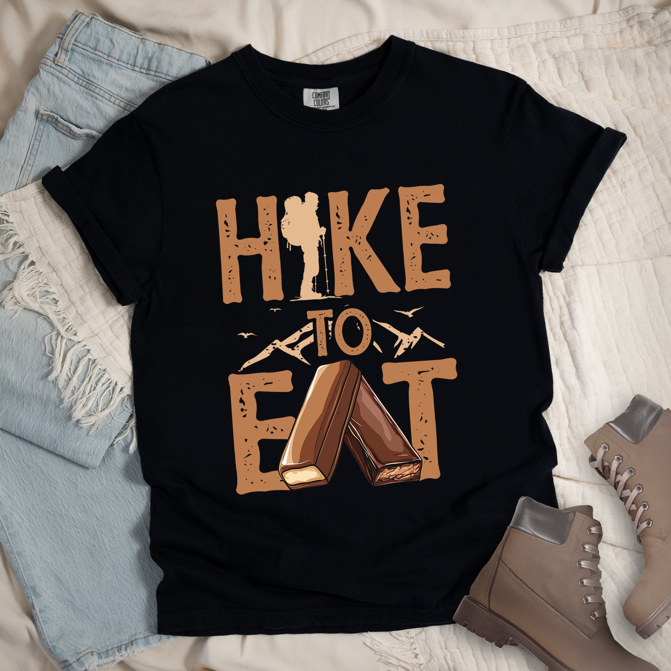 Black t-shirt with text "HIKE TO EAT". "HIKE" appears in large brown letters at the top with a hiker silhouette integrated into the letter "I", "TO" in brown text with small mountain outlines on either side in the middle, and "EAT" in large brown letters at the bottom with two chocolate candy bars arranged in an "A" shape replacing the letter "A". Small flying birds appear near "TO". The shirt features a classic crew neck and casual rolled sleeves, styled alongside light wash denim and brown hiking boots.