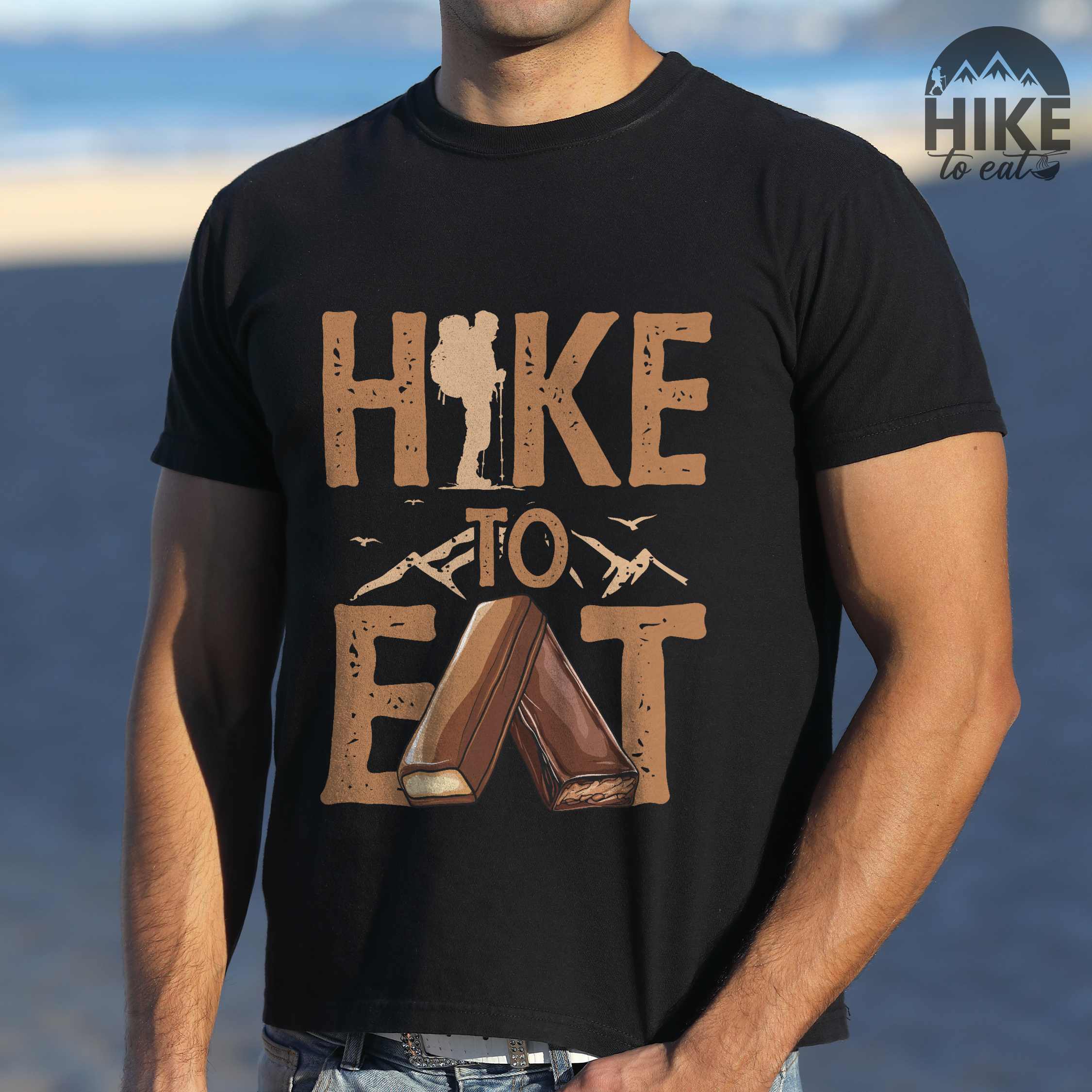 Man wearing black "Hike to Eat" colored statement tee with bold typography, mountain, illustrated bars of chocolate, perfect for chocolate lovers.