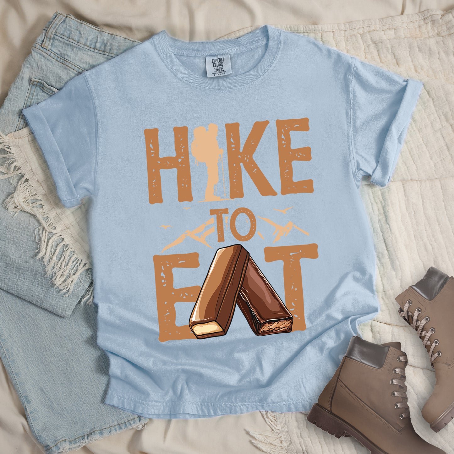 Chambray (Light Blue) t-shirt with text "HIKE TO EAT". "HIKE" appears in large brown letters at the top with a hiker silhouette integrated into the letter "I", "TO" in brown text with small mountain outlines on either side in the middle, and "EAT" in large brown letters at the bottom with two chocolate candy bars arranged in an "A" shape replacing the letter "A". Small flying birds appear near "TO". Features a classic crew neck and casual rolled sleeves, alongside light wash denim and brown hiking boots.