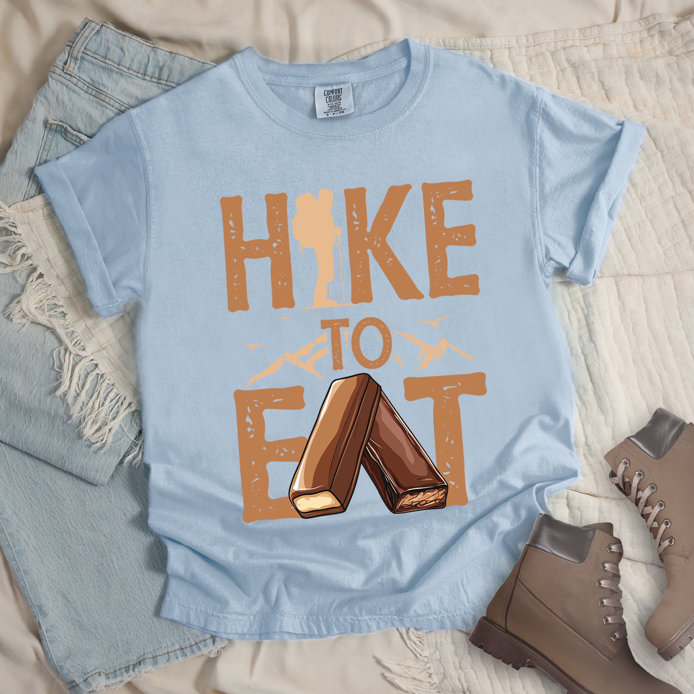 Chambray (Light Blue) t-shirt with text "HIKE TO EAT". "HIKE" appears in large brown letters at the top with a hiker silhouette integrated into the letter "I", "TO" in brown text with small mountain outlines on either side in the middle, and "EAT" in large brown letters at the bottom with two chocolate candy bars arranged in an "A" shape replacing the letter "A". Small flying birds appear near "TO". Features a classic crew neck and casual rolled sleeves, alongside light wash denim and brown hiking boots.
