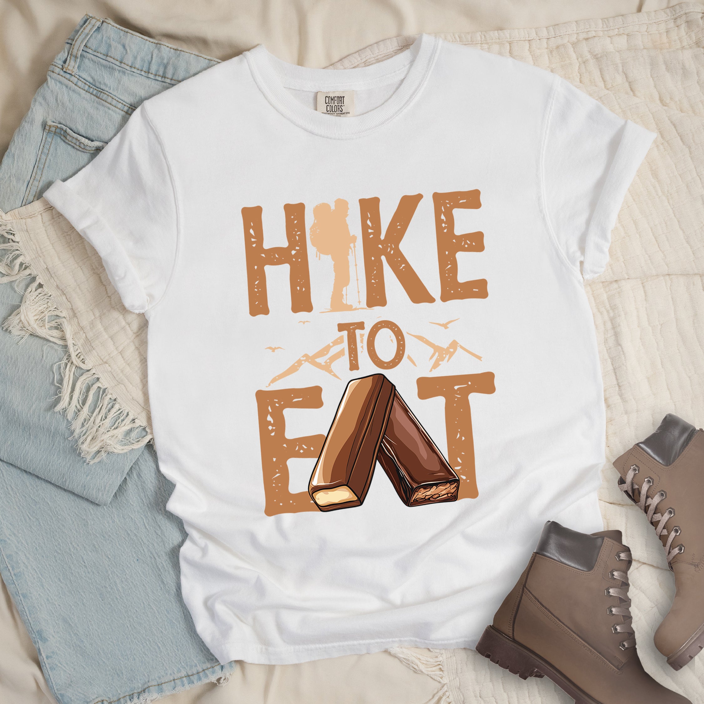 White t-shirt with text "HIKE TO EAT". "HIKE" appears in large brown letters at the top with a hiker silhouette integrated into the letter "I", "TO" in brown text with small mountain outlines on either side in the middle, and "EAT" in large brown letters at the bottom with two chocolate candy bars arranged in an "A" shape replacing the letter "A". Small flying birds appear near "TO". The shirt features a classic crew neck and casual rolled sleeves, styled alongside light wash denim and brown hiking boots.