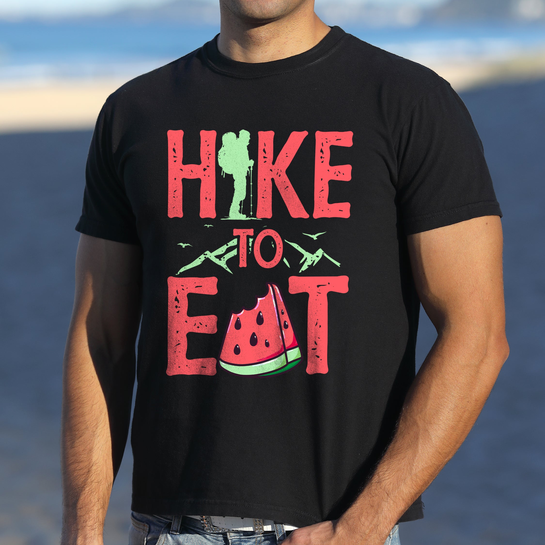 Black graphic t-shirt with 'Hike to Eat' text in red lettering featuring green hiker silhouette, mountain line art, and watermelon slice illustration.