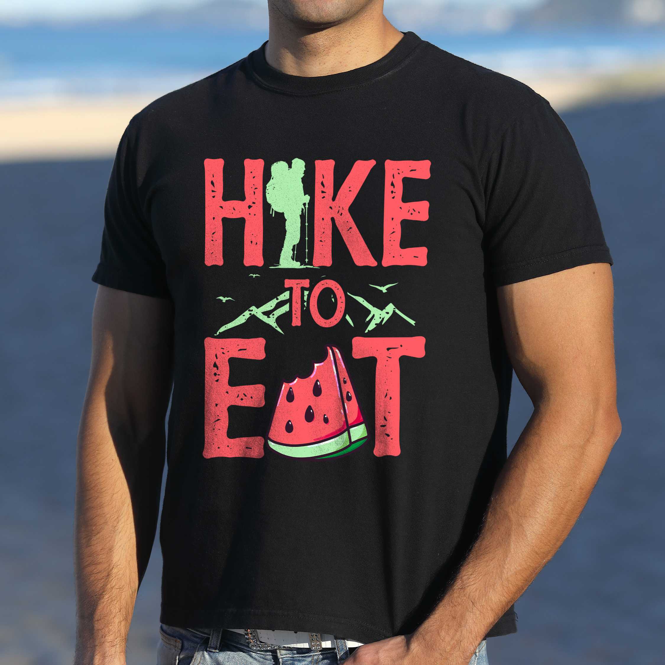 Man wearing black "Hike to Eat" colored statement tee with bold typography, mountain, illustrated slice of watermelon, perfect for fruit lovers.