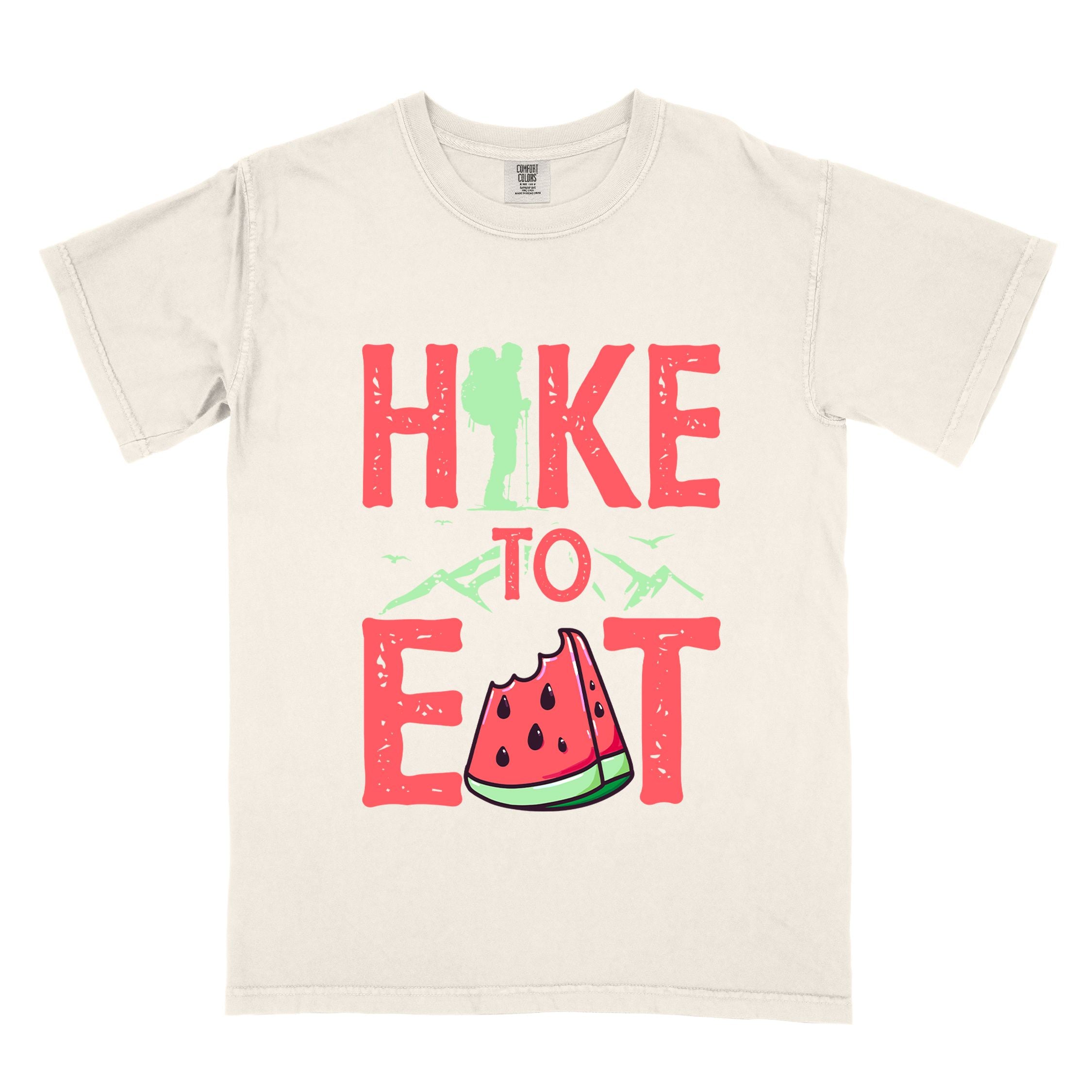 Ivory "Hike to Eat" colored statement tee with bold typography, mountain, illustrated slice of watermelon, perfect for fruit lovers.