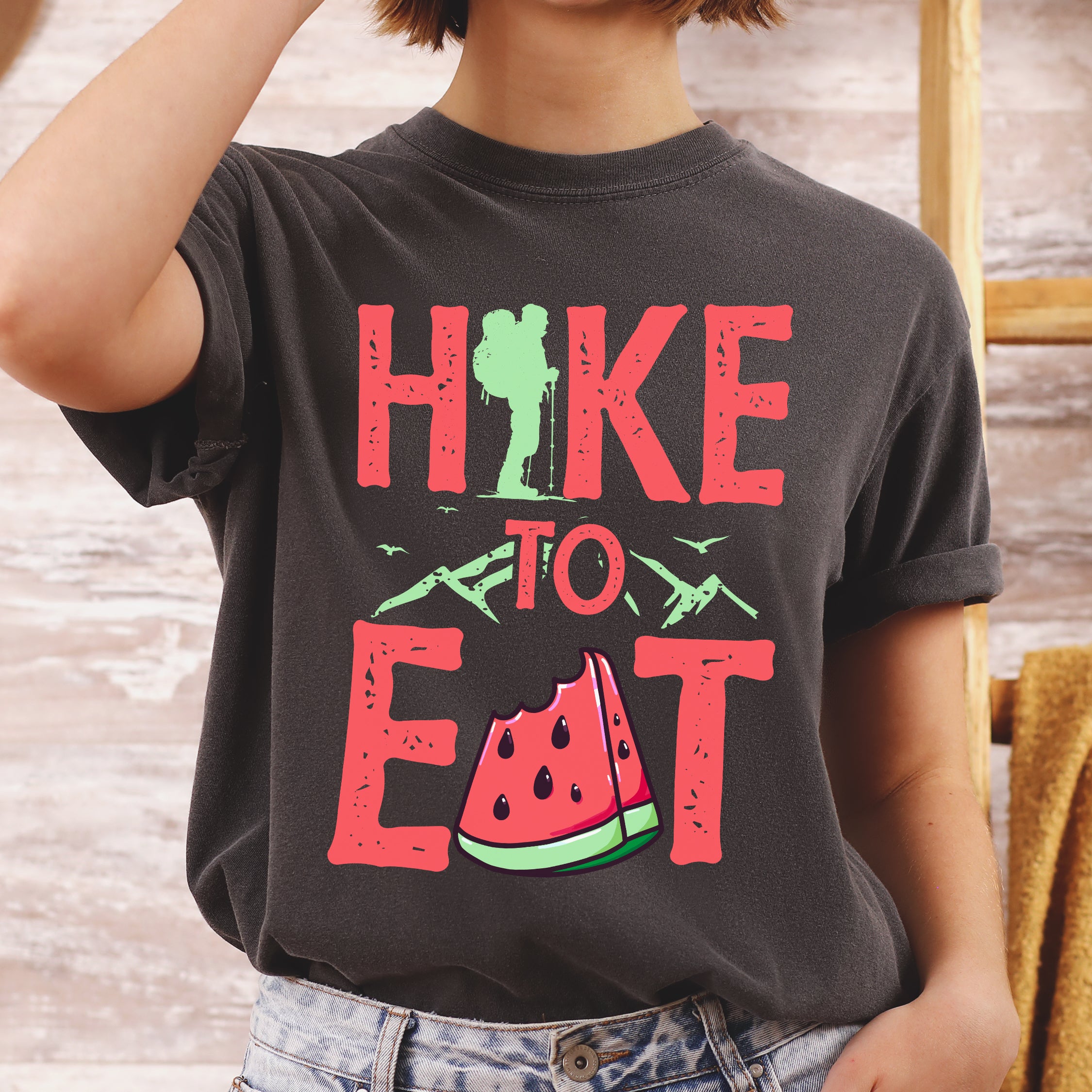 Pepper (Dark Gray) graphic t-shirt with 'Hike to Eat' text in red lettering featuring green hiker silhouette, mountain line art, and watermelon slice illustration