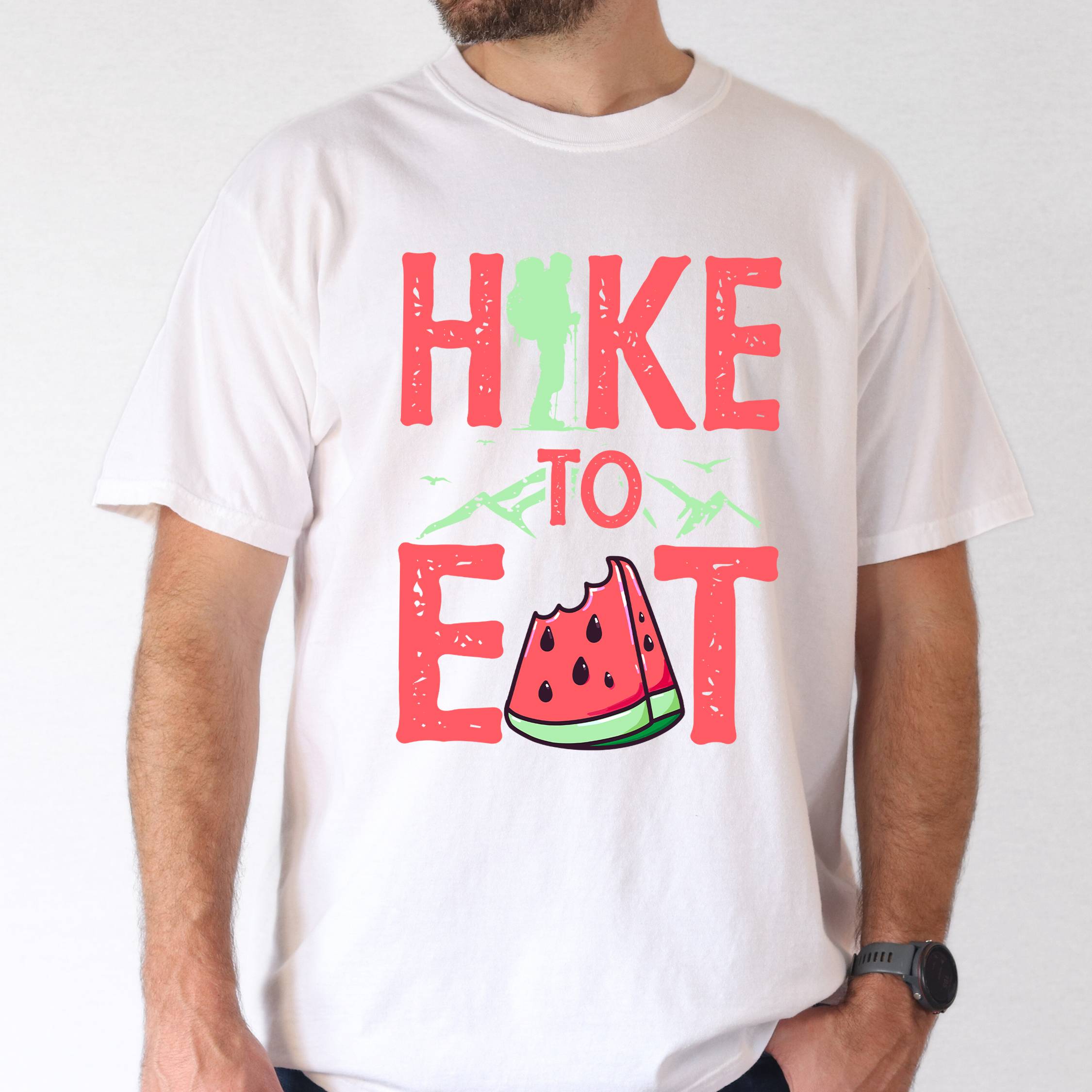 Man wearing white "Hike to Eat" colored statement tee with bold typography, mountain, illustrated slice of watermelon, perfect for fruit lovers.