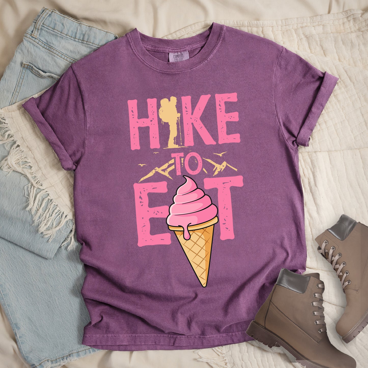 Berry (Dark Violet) t-shirt with text "HIKE TO EAT". "HIKE" appears in large pink letters at the top with a yellow hiker silhouette integrated into the letter "I", "TO" in pink text with yellow mountain outlines on either side in the middle, and "EAT" in large pink letters at the bottom with a pink ice cream cone in a waffle cone positioned between "E" and "T". Small flying birds appear near "TO". Features a classic crew neck and casual rolled sleeves, alongside light wash denim and brown hiking boots.