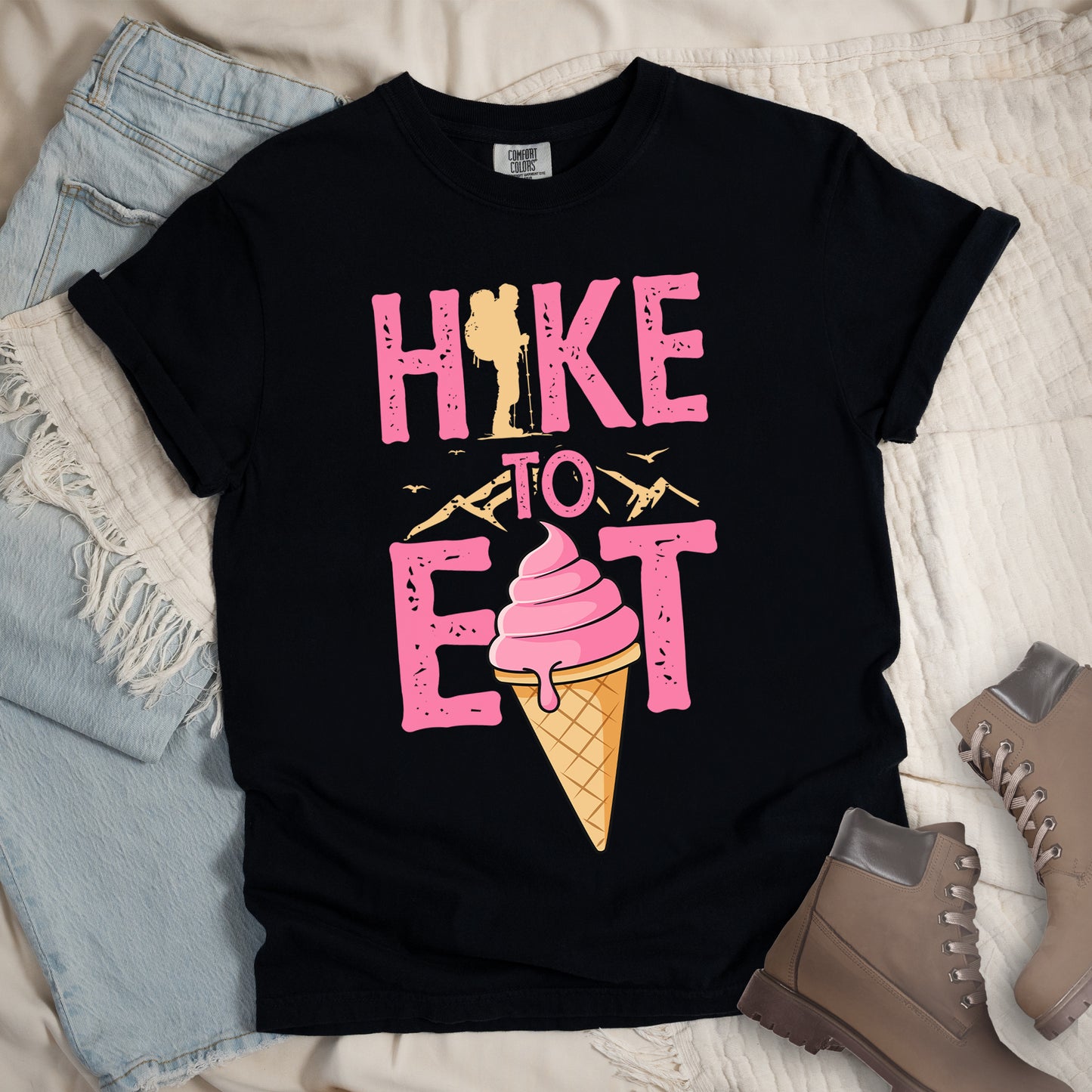 Black t-shirt with text reading "HIKE TO EAT". "HIKE" appears in large pink letters at the top with a yellow hiker silhouette integrated into the letter "I", "TO" in pink text with yellow mountain outlines on either side in the middle, and "EAT" in large pink letters at the bottom with a pink ice cream cone in a waffle cone positioned between "E" and "T". Small flying birds appear near "TO". Features a classic crew neck and casual rolled sleeves, styled alongside light wash denim and brown hiking boots.