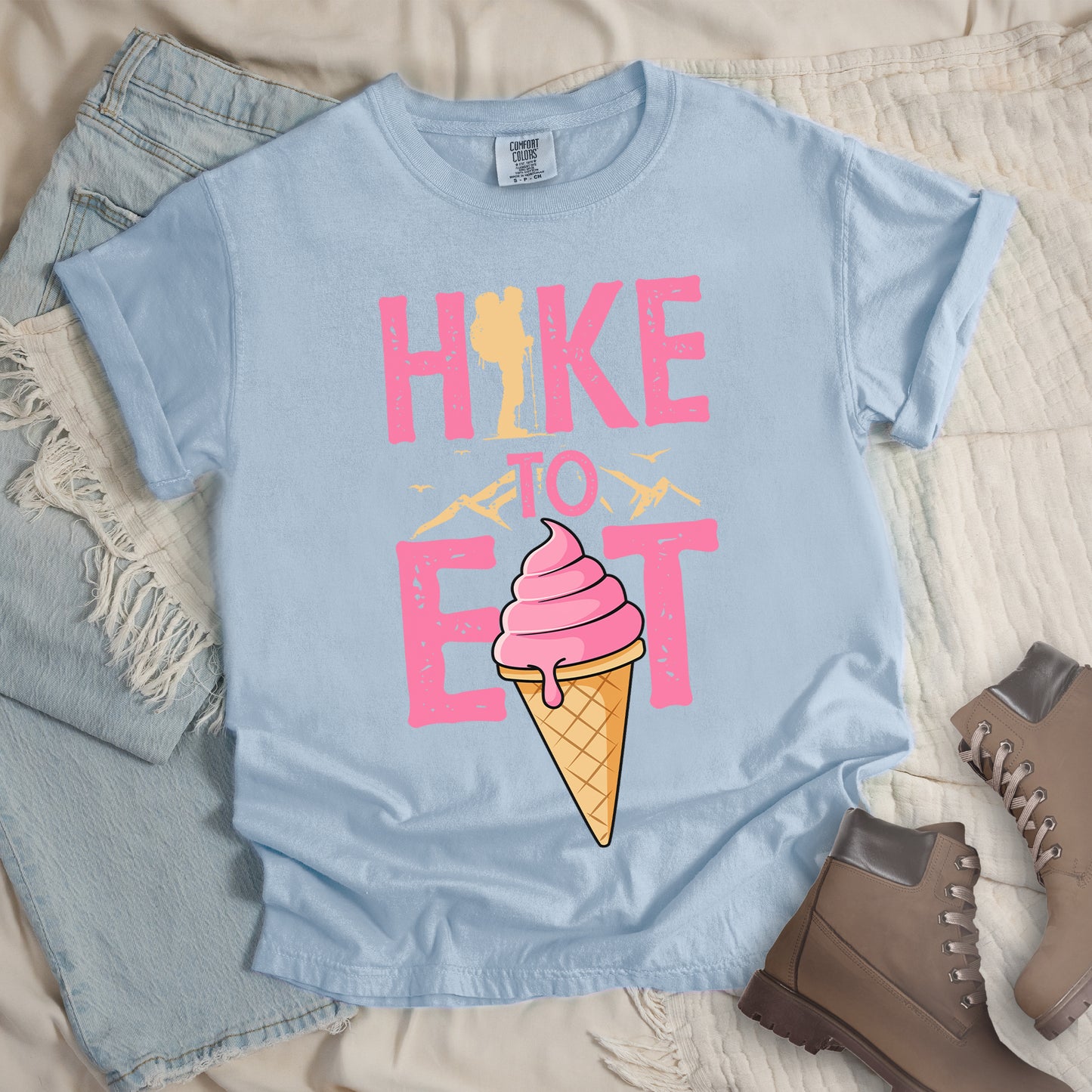 Chambray (Light Blue) t-shirt with text "HIKE TO EAT". "HIKE" appears in large pink letters at the top with a yellow hiker silhouette integrated into the letter "I", "TO" in pink text with yellow mountain outlines on either side in the middle, and "EAT" in large pink letters at the bottom with a pink ice cream cone in a waffle cone positioned between "E" and "T". Small flying birds appear near "TO". Features a classic crew neck and casual rolled sleeves, alongside light wash denim and brown hiking boots.