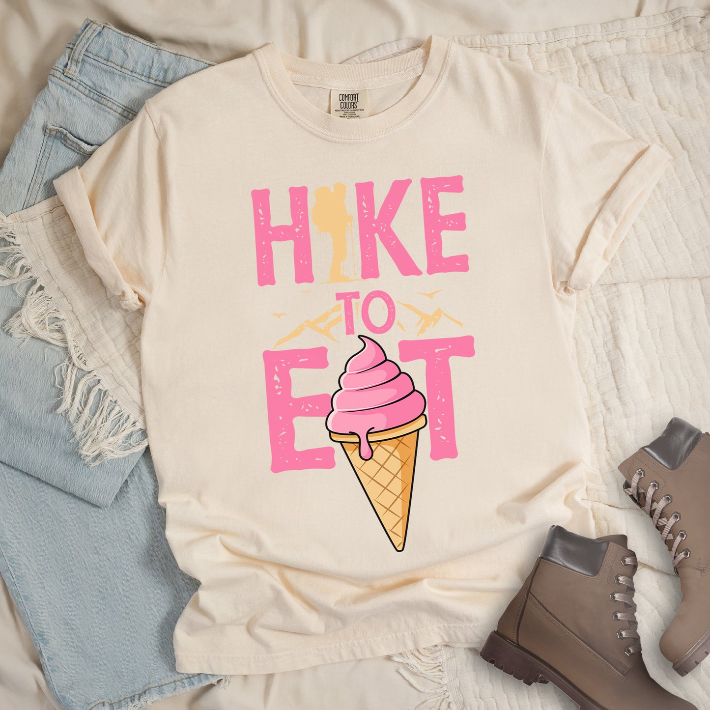 Ivory (Cream White) t-shirt with text "HIKE TO EAT". "HIKE" appears in large pink letters at the top with a yellow hiker silhouette integrated into the letter "I", "TO" in pink text with yellow mountain outlines on either side in the middle, and "EAT" in large pink letters at the bottom with a pink ice cream cone in a waffle cone positioned between "E" and "T". Small flying birds appear near "TO". Features a classic crew neck and casual rolled sleeves, alongside light wash denim and brown hiking boots.