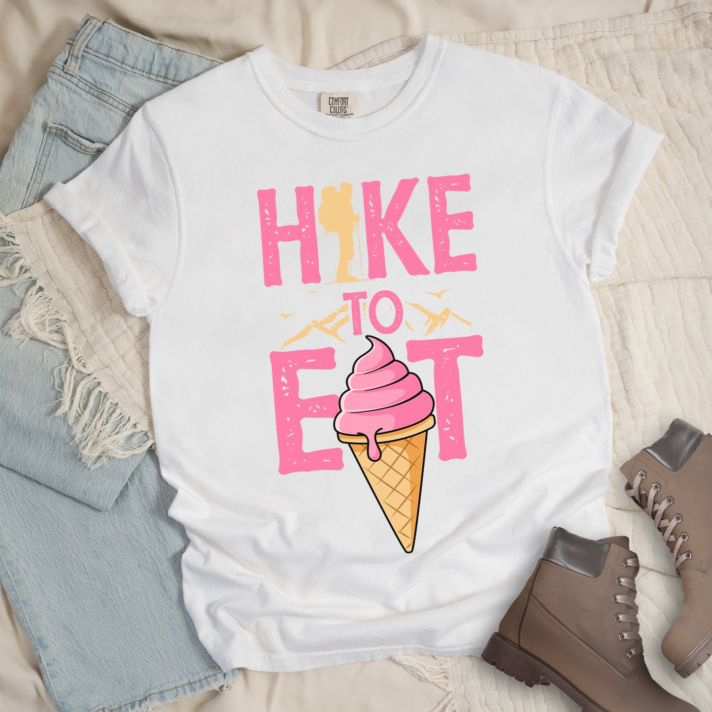 White t-shirt with text reading "HIKE TO EAT". "HIKE" appears in large pink letters at the top with a yellow hiker silhouette integrated into the letter "I", "TO" in pink text with yellow mountain outlines on either side in the middle, and "EAT" in large pink letters at the bottom with a pink ice cream cone in a waffle cone positioned between "E" and "T". Small flying birds appear near "TO". Features a classic crew neck and casual rolled sleeves, styled alongside light wash denim and brown hiking boots.