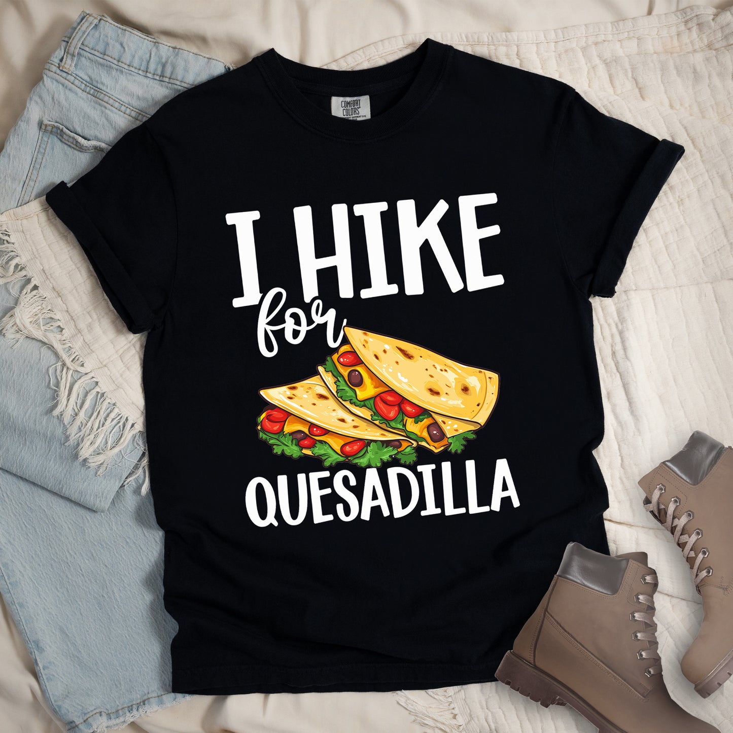 Black t-shirt with white text reading "I HIKE for QUESADILLA". The words "I HIKE" appear at the top in large capital letters, "for" in cursive script in the middle, and "QUESADILLA" at the bottom in capital letters. Between "for" and "QUESADILLA" is a colorful illustration of quesadillas filled with cheese, tomatoes, and lettuce. Features a classic crew neck and casual rolled sleeves, styled alongside light wash denim and brown hiking boots.