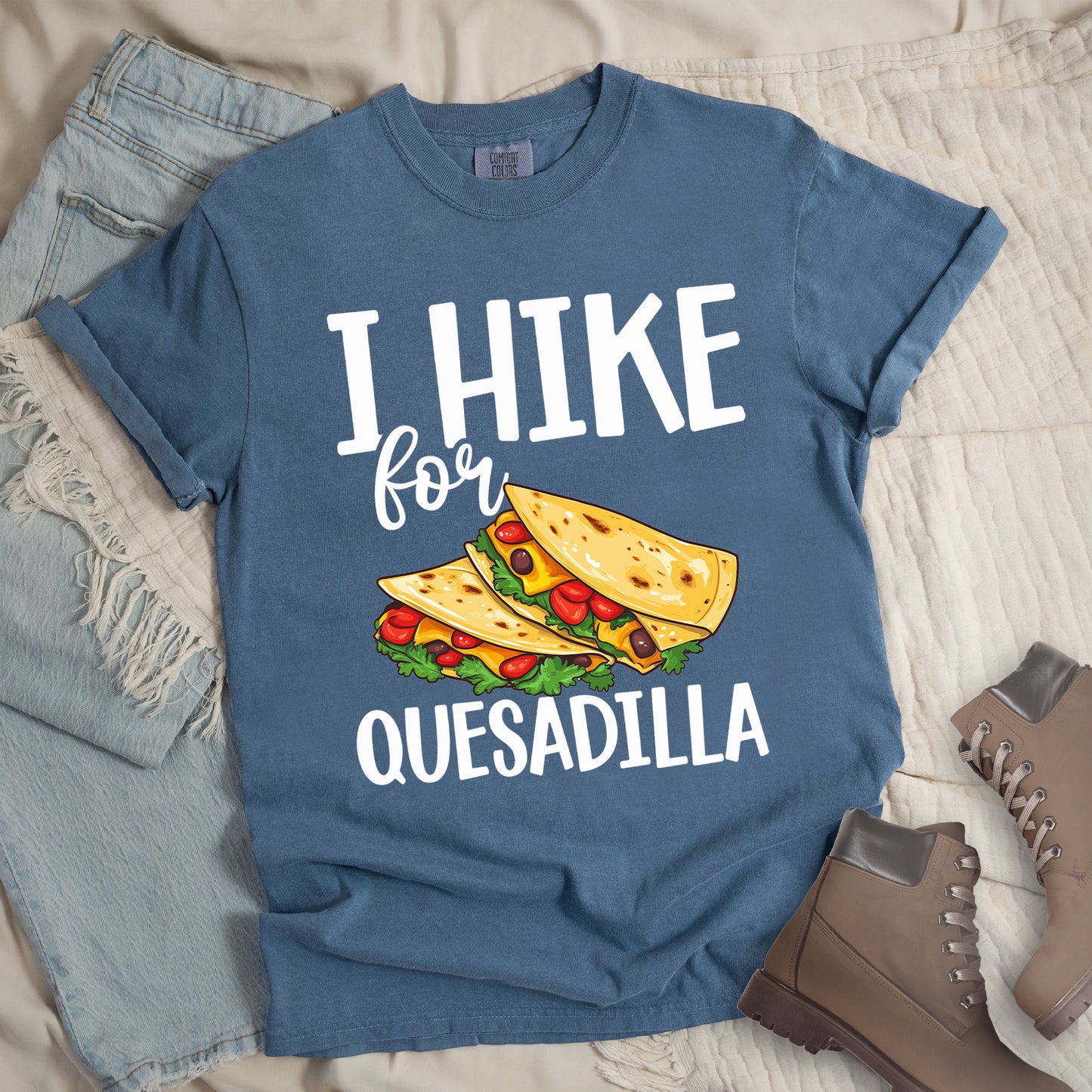 Blue Jean (Denim Blue) t-shirt with white text reading "I HIKE for QUESADILLA". The words "I HIKE" appear at the top in large capital letters, "for" in cursive script in the middle, and "QUESADILLA" at the bottom in capital letters. Between "for" and "QUESADILLA" is a colorful illustration of quesadillas filled with cheese, tomatoes, and lettuce. Features a classic crew neck and casual rolled sleeves, styled alongside light wash denim and brown hiking boots.
