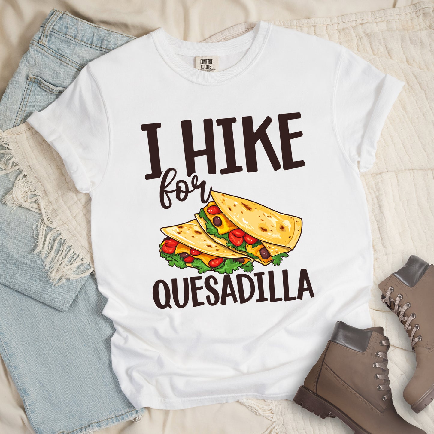 White t-shirt with dark brown text reading "I HIKE for QUESADILLA". The words "I HIKE" appear at the top in large capital letters, "for" in cursive script in the middle, and "QUESADILLA" at the bottom in capital letters. Between "for" and "QUESADILLA" is a colorful illustration of quesadillas filled with cheese, tomatoes, and lettuce. Features a classic crew neck and casual rolled sleeves, styled alongside light wash denim and brown hiking boots.