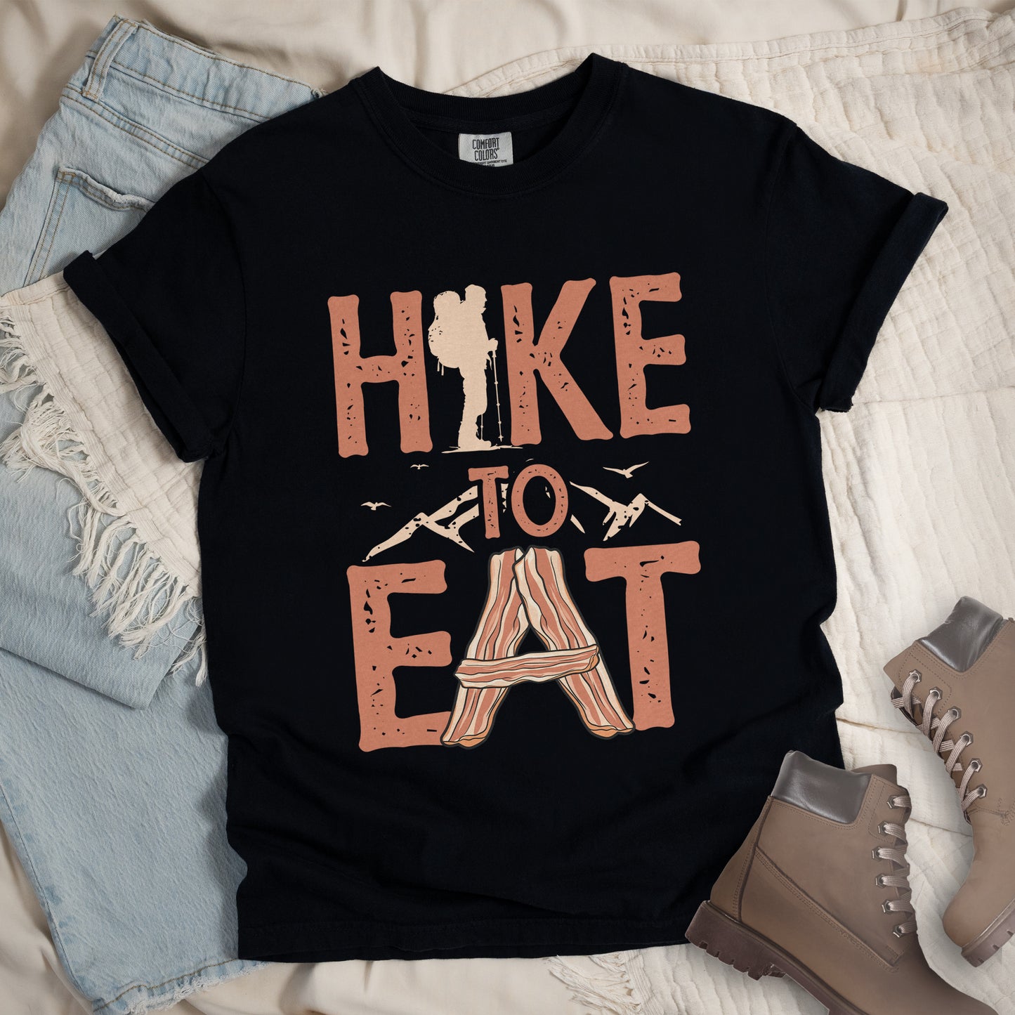 Black t-shirt with text reading "HIKE TO EAT". "HIKE" appears in large orange letters at the top with a beige hiker silhouette integrated into the letter "I", "TO" in orange text with mountain outlines on either side in the middle, and "EAT" in large orange letters at the bottom with strips of bacon arranged in an "A" shape replacing the letter "A". Small flying birds appear near "TO". The shirt features a classic crew neck and casual rolled sleeves, styled alongside light wash denim and brown hiking boots