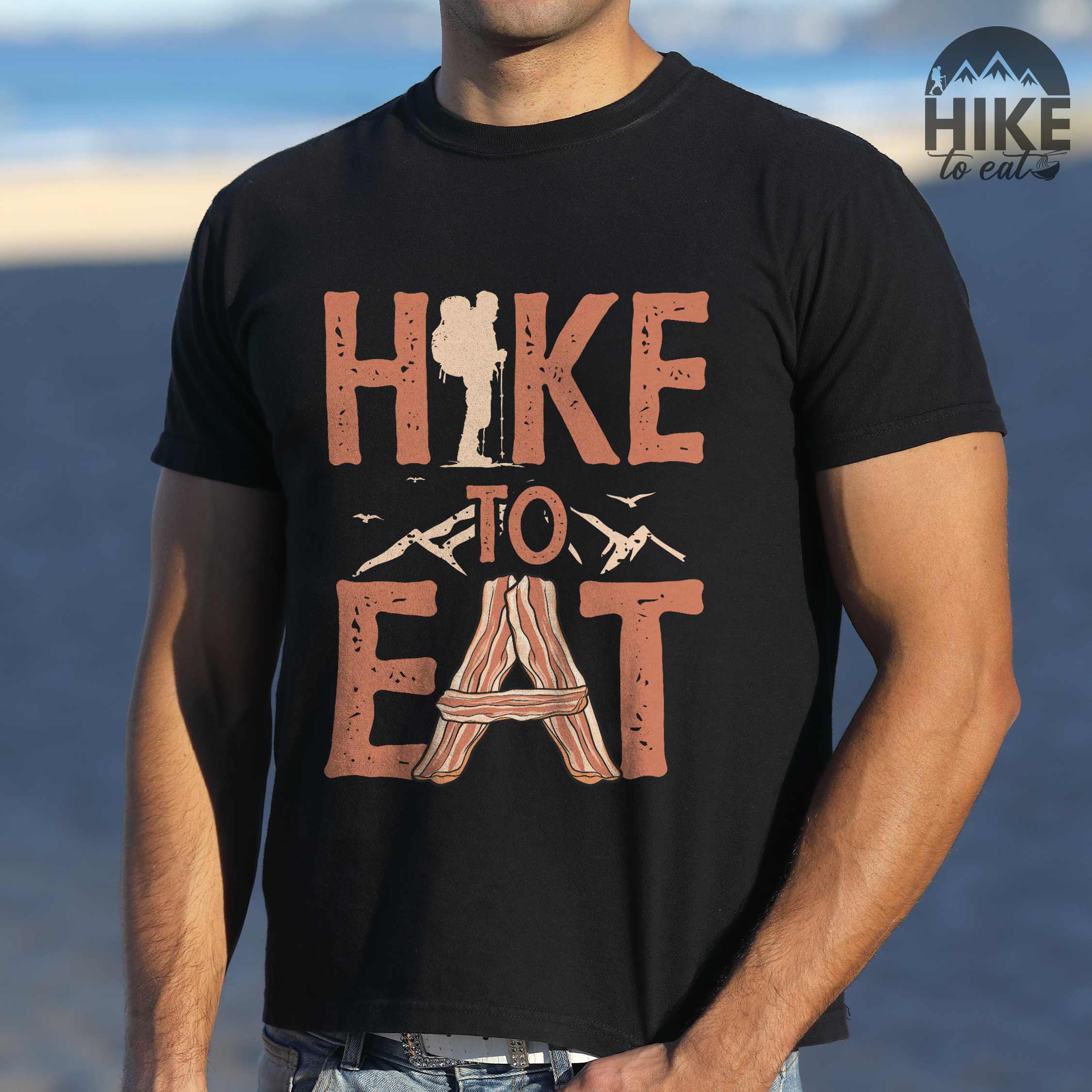 man wearing black"Hike to Eat" colored statement tee with bold typography, mountain, illustrated slices of bacon, and backpacker for breakfast enthusiasts.