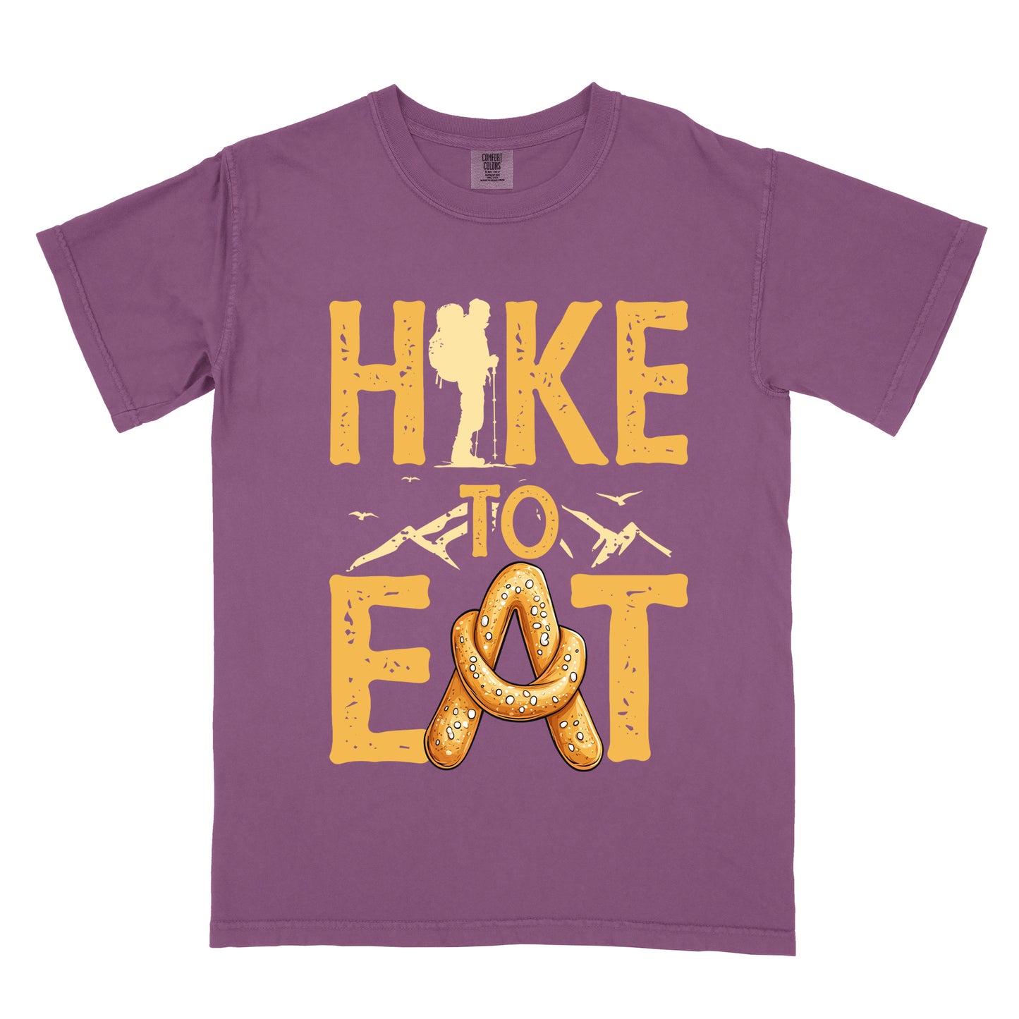 Berry (Dark Violet) t-shirt with golden yellow distressed text reading "HIKE TO EAT". "HIKE" appears in large golden yellow letters at the top with a hiker silhouette integrated into the letter "I", "TO" in golden yellow text with mountain outlines on either side in the middle, and "EAT" in large golden yellow letters at the bottom with a pretzel shaped in an "A" replacing the letter "A". Small flying birds appear near "TO".