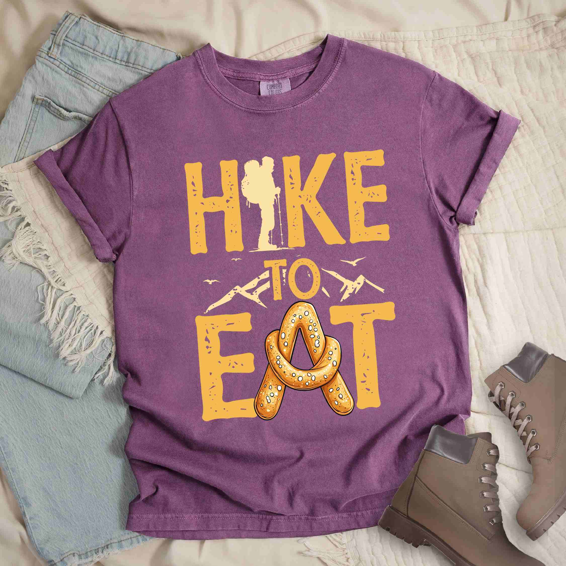 Berry "Hike to Eat" colored statement tee with bold typography, mountain, illustrated baked pretzel, and backpacker for pastry enthusiasts.
