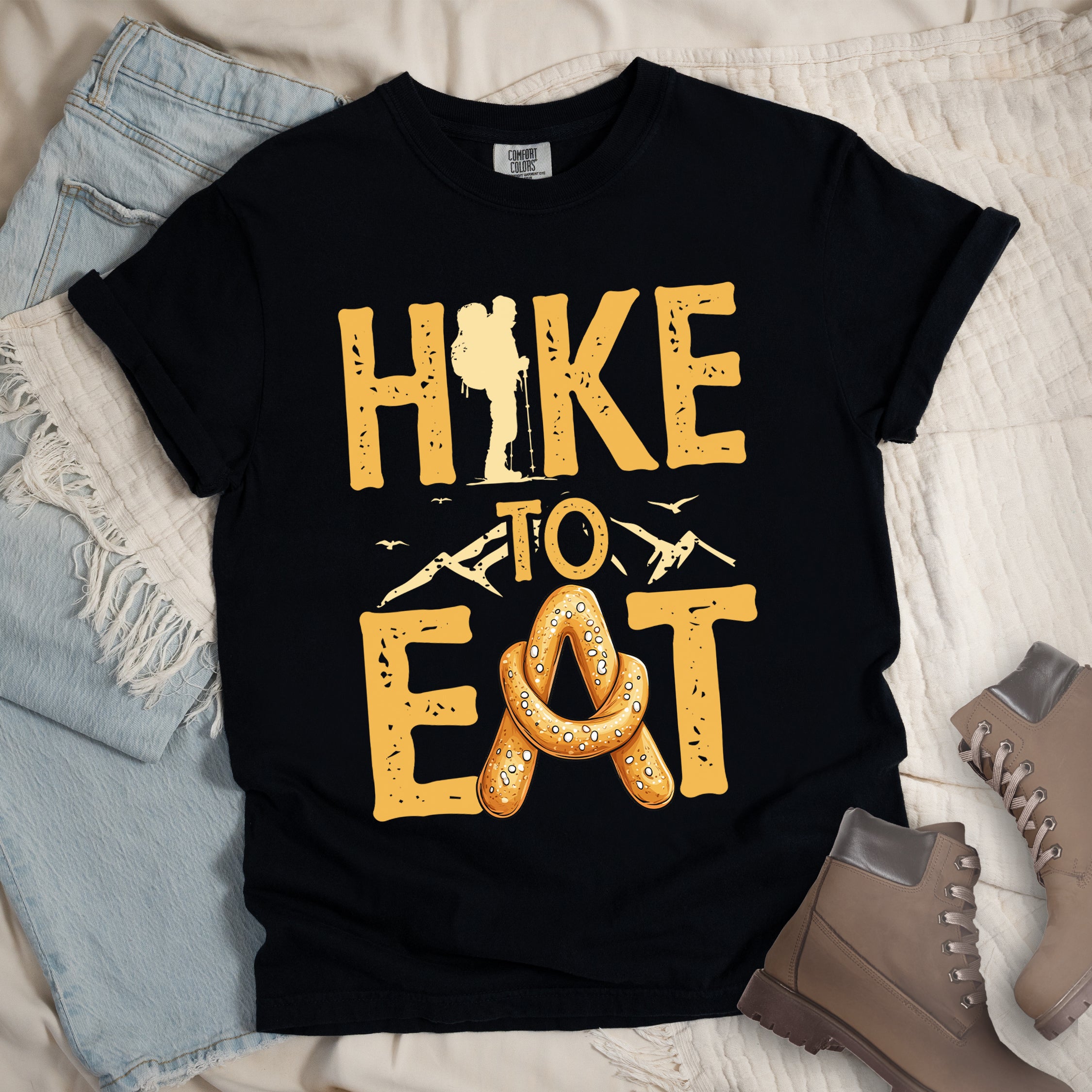 Black Light Green t-shirt with text reading "HIKE TO EAT". "HIKE" appears in golden yellow letters at the top with a hiker silhouette integrated into the letter "I", "TO" in golden yellow text with mountain outlines on either side in the middle, and "EAT" in large golden yellow letters at the bottom with a pretzel shaped in an "A" replacing the letter "A". Small flying birds appear near "TO". Features a classic crew neck and casual rolled sleeves, styled alongside light wash denim and brown hiking boots.