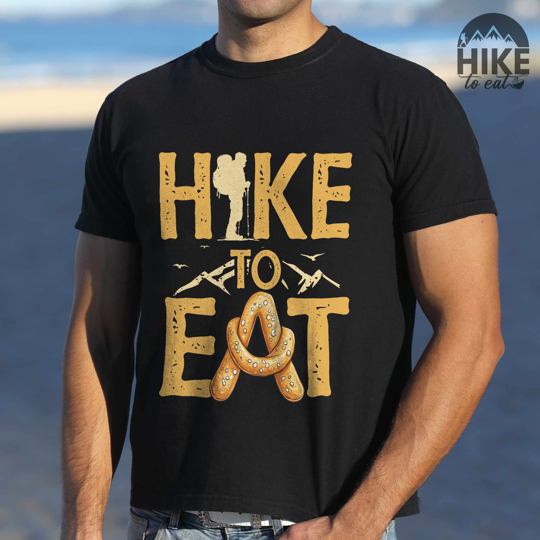 Man wearing black "Hike to Eat" colored statement tee with bold typography, mountain, illustrated baked pretzel, and backpacker for pastry enthusiasts.